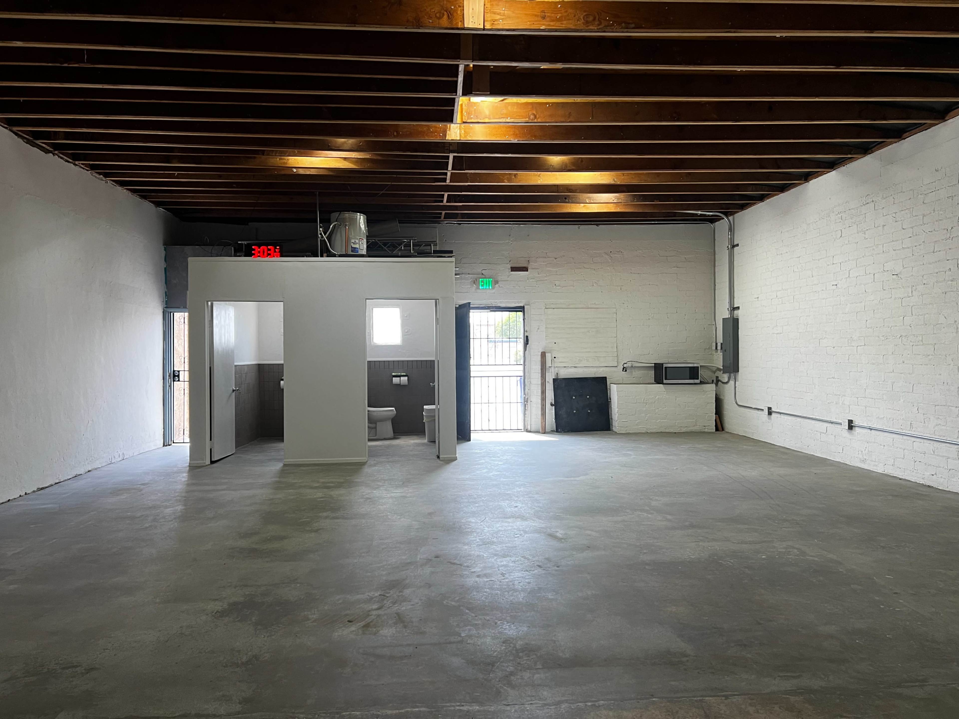 An empty room with exposed wooden beams, concrete flooring, and a small restroom enclosed in a partition.