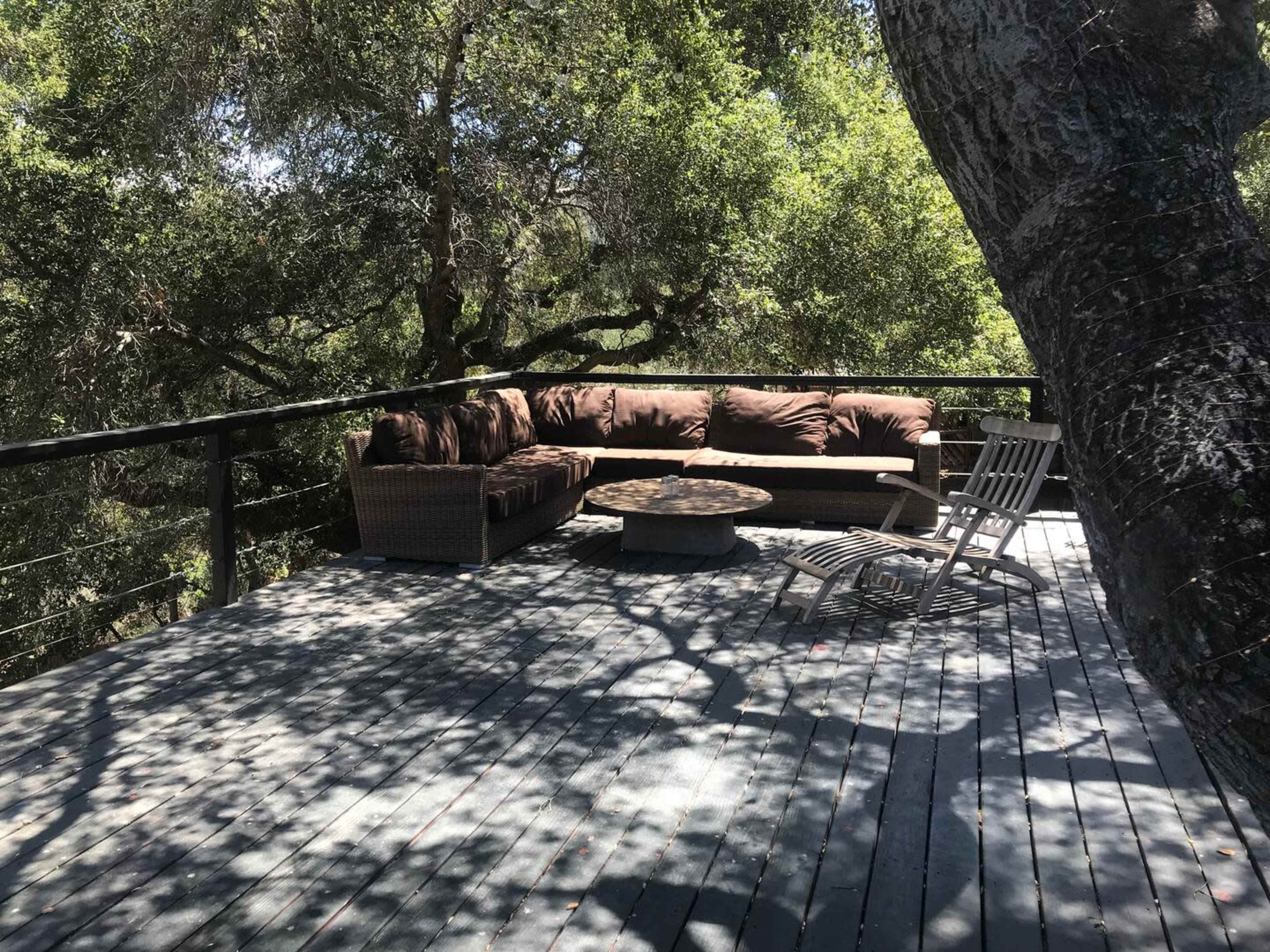 A shaded outdoor deck features a brown sectional couch, a circular stone table, and a wooden lounge chair surrounded by trees.