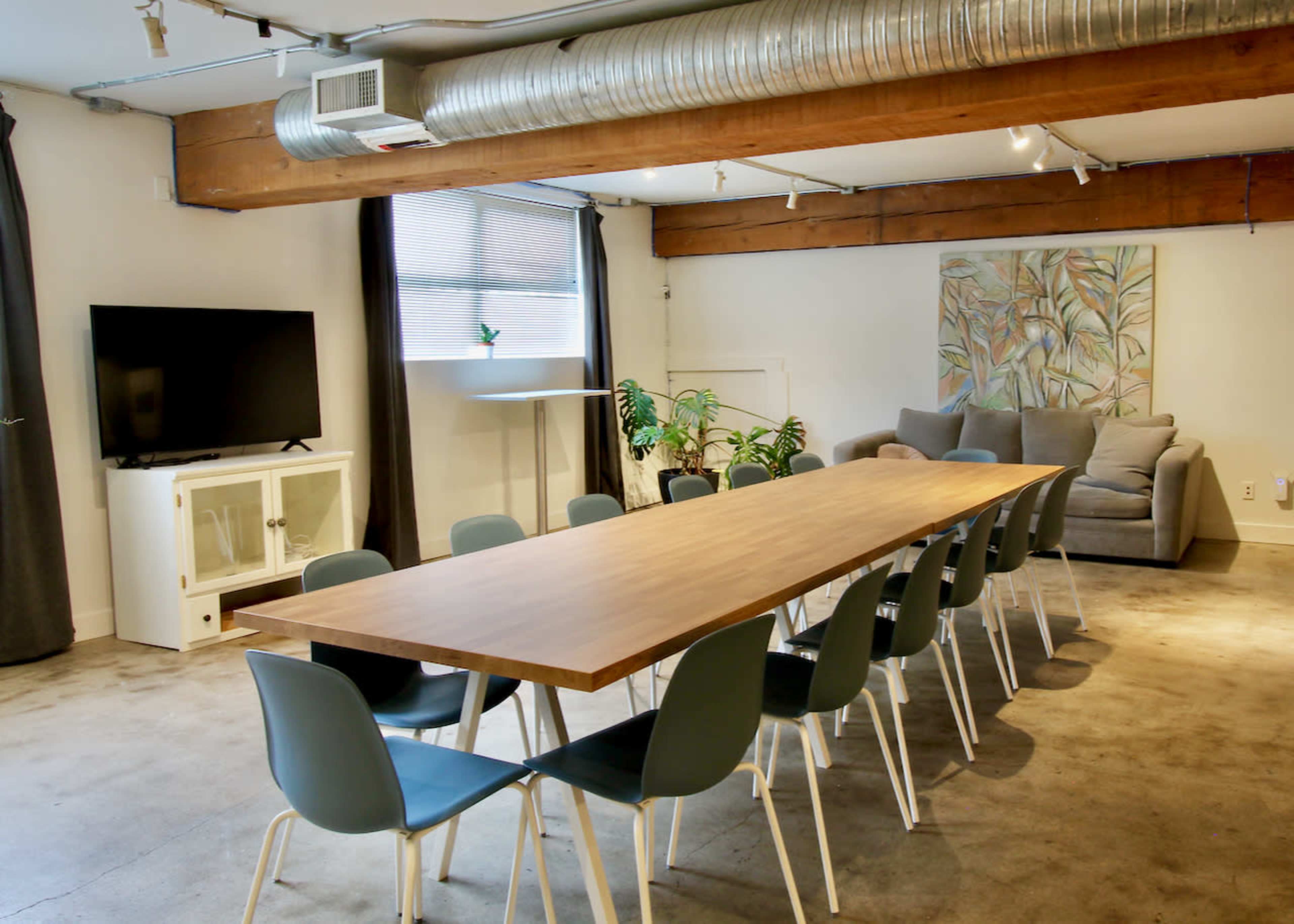 The image features a spacious conference room with a long wooden table surrounded by chairs, a television mounted on the wall, and a gray couch in the corner.