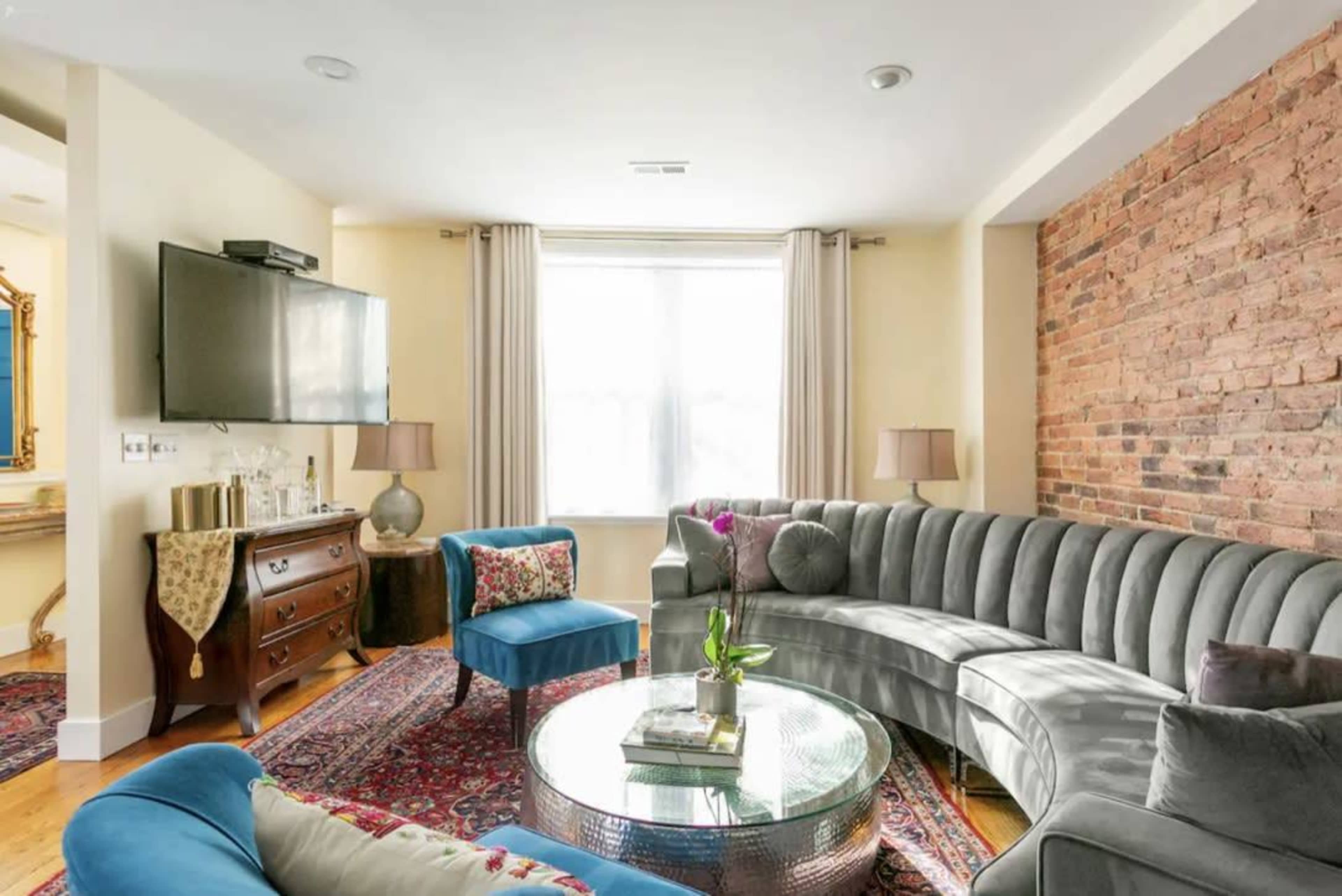 The living room features a curved gray sofa, a round glass coffee table, and a combination of colorful seating, with a window illuminating the space.
