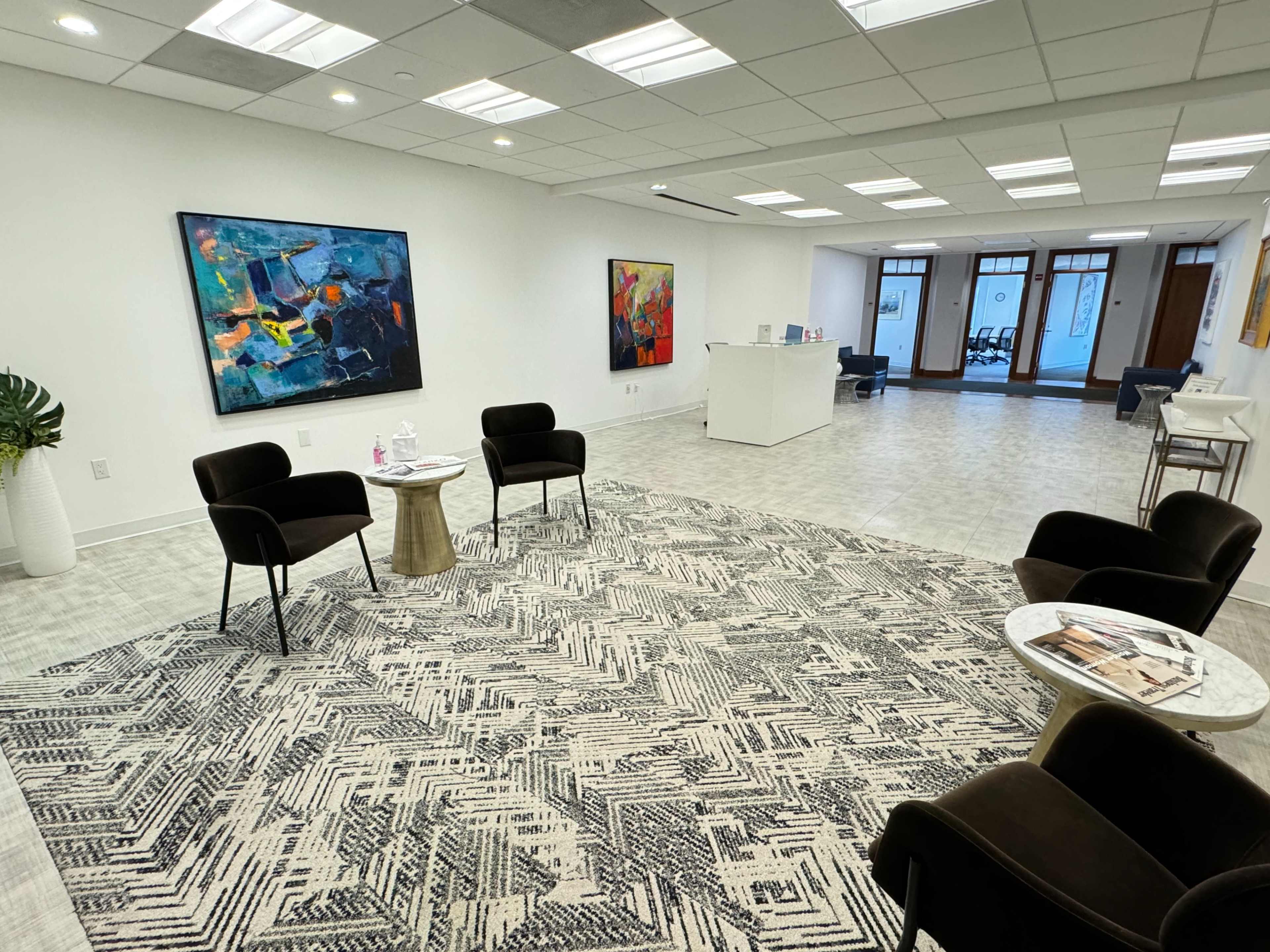 The image shows a modern office waiting area with two black chairs and a round table on a patterned rug, surrounded by abstract art on the walls and a reception desk in the background.