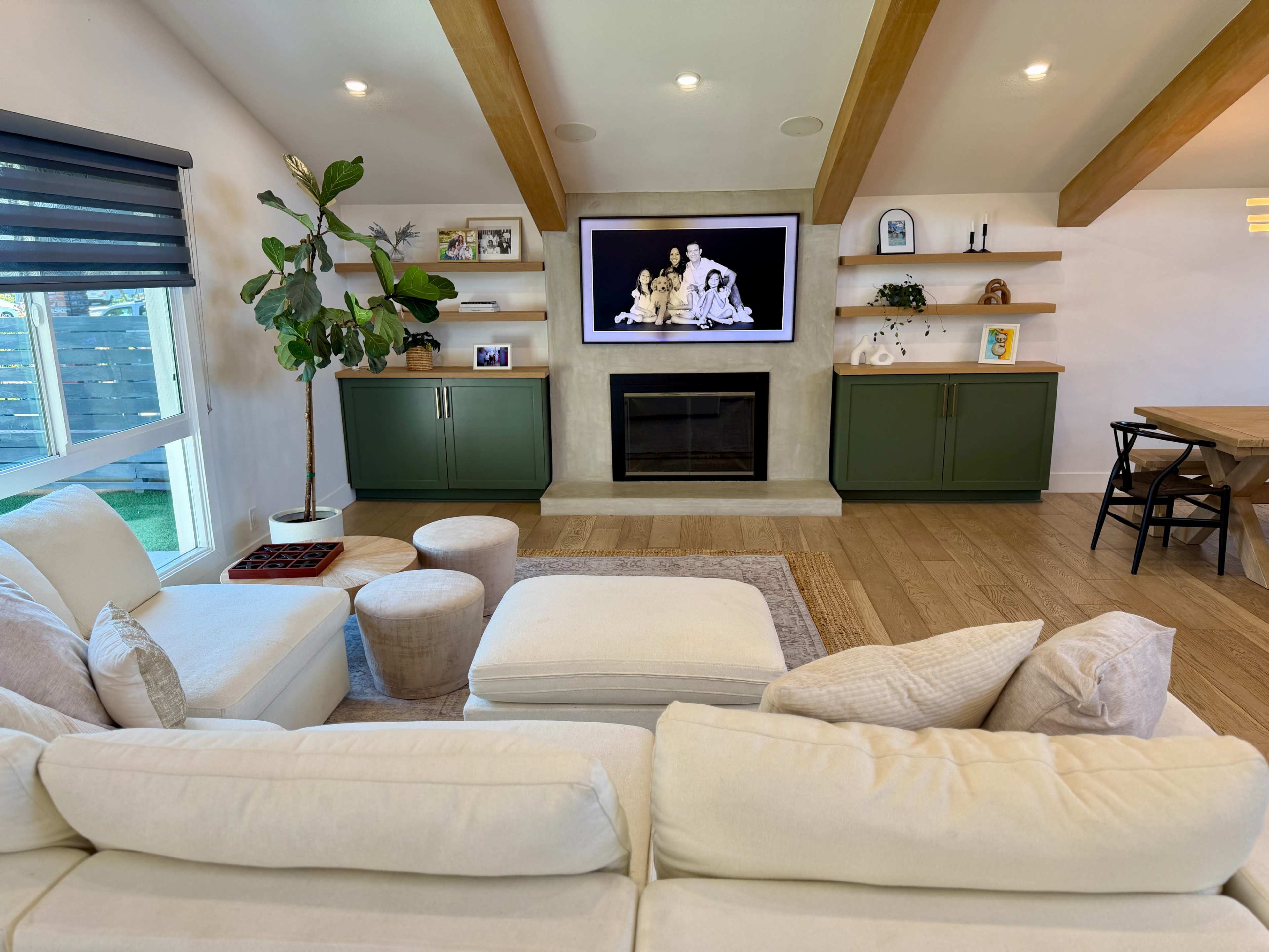 The image shows a modern living room featuring a white sectional sofa, a large flat-screen TV mounted above a fireplace, built-in bookshelves, and a dining area with a wooden table.
