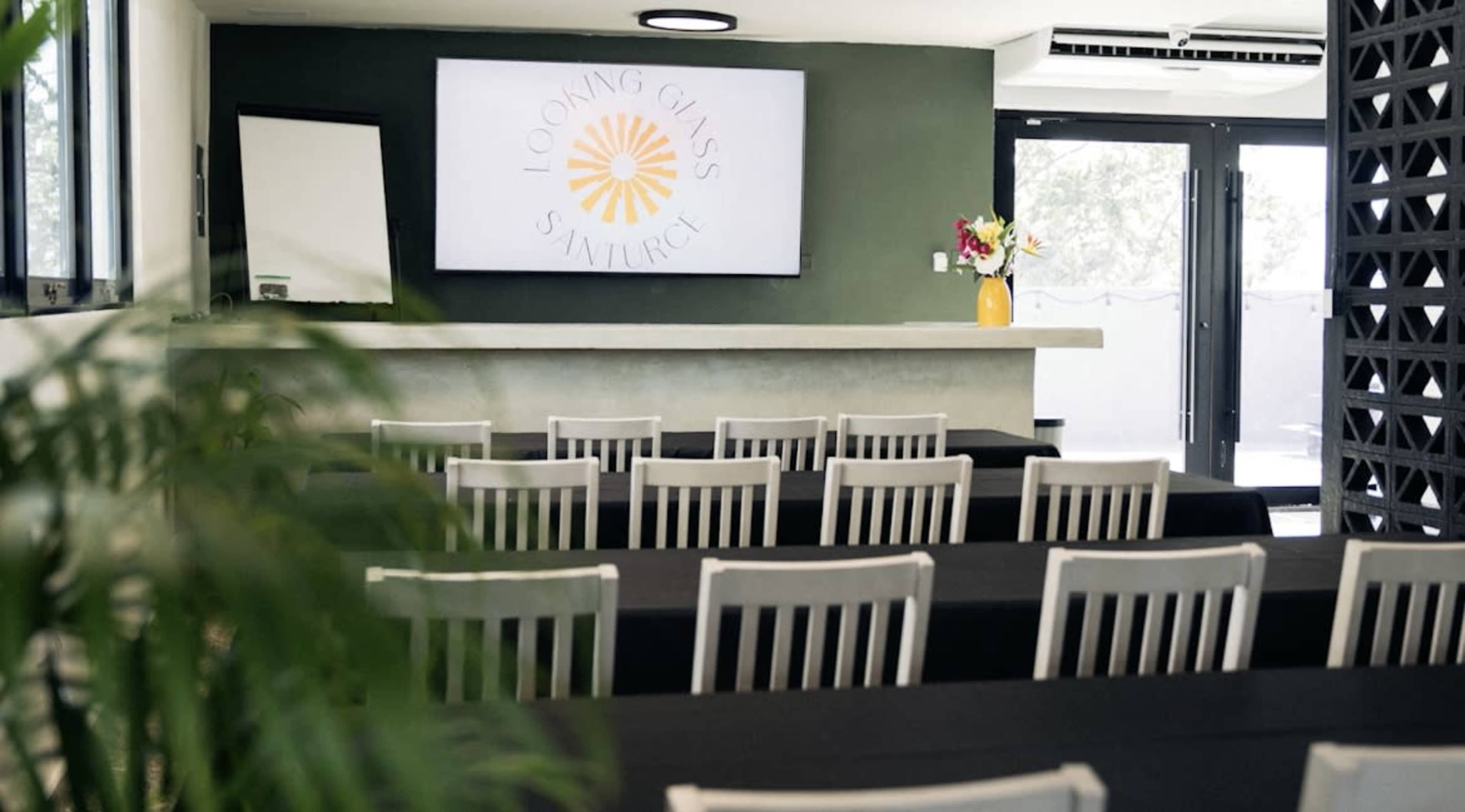 The image shows a seminar room with rows of white chairs facing a large screen displaying a logo, alongside a counter with a vase of flowers.