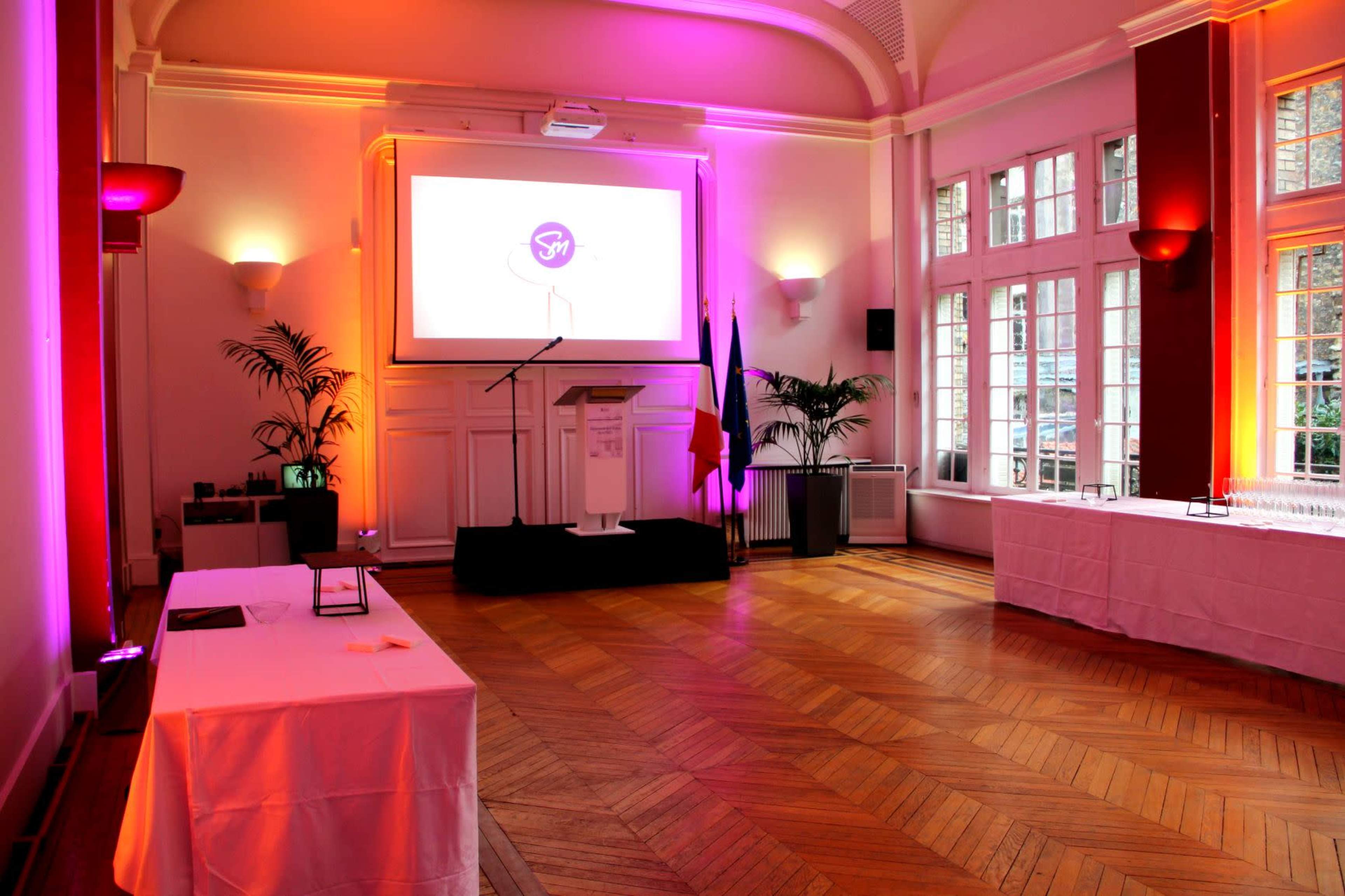 A spacious room is set up for an event, featuring a podium, a large screen, and tables with white linens, illuminated by colorful lighting.