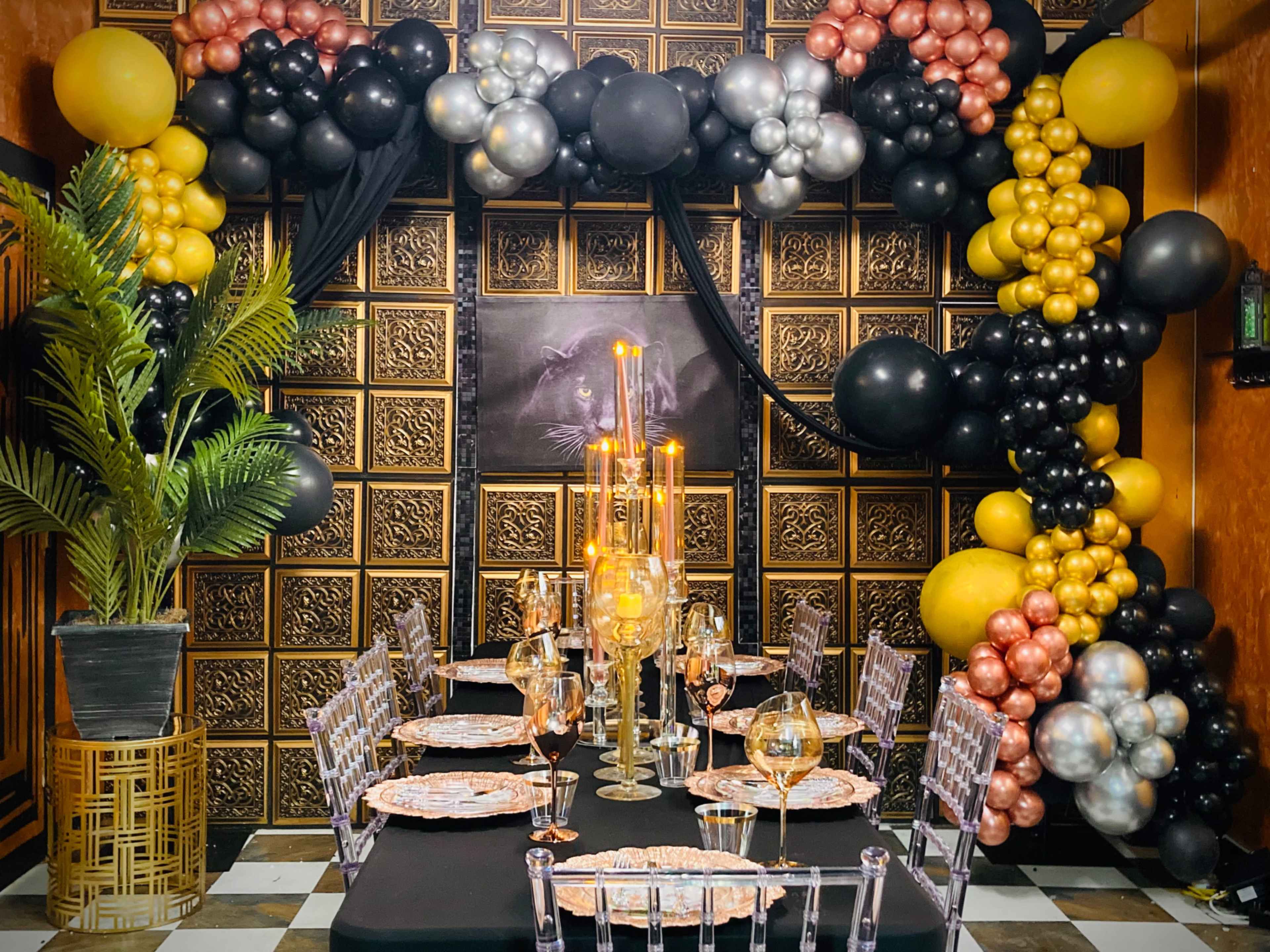 A decorated dining area with a table set for a formal meal, surrounded by a balloon arch in black, gold, and silver, and featuring a large plant in a decorative pot.