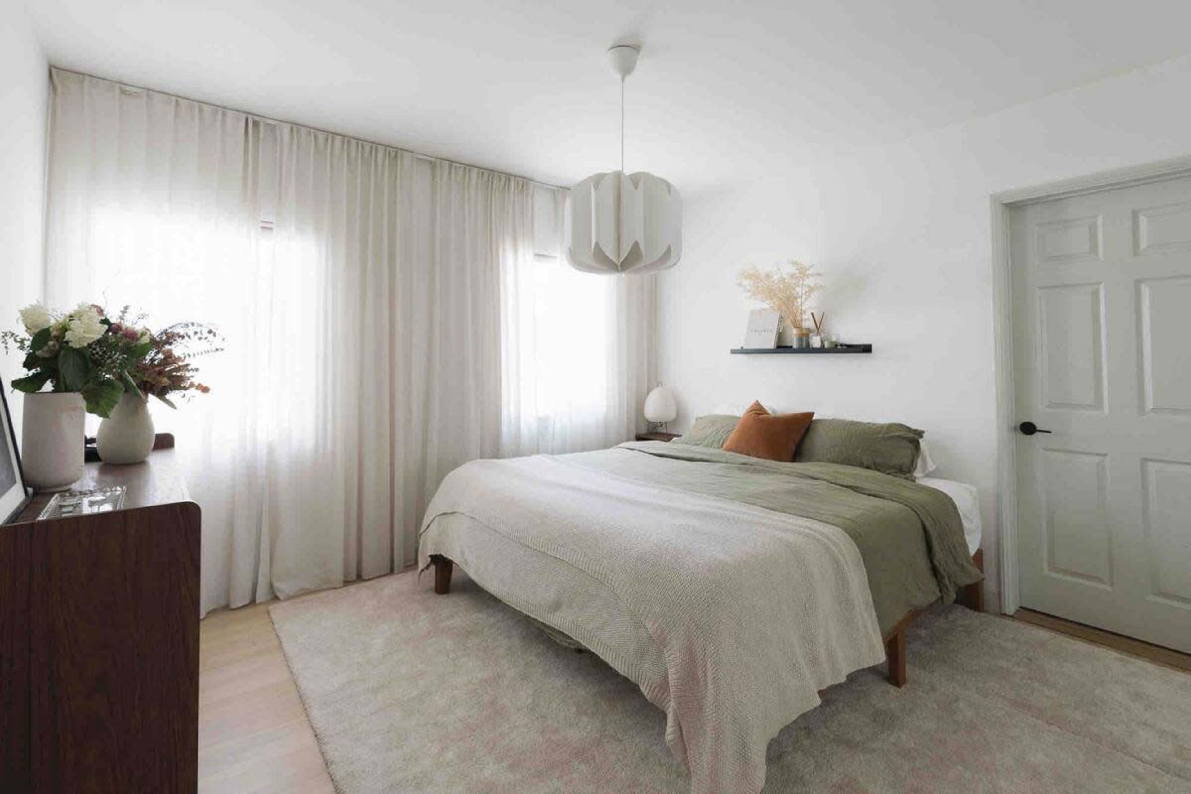 A neatly arranged bedroom features a large bed with green and neutral bedding, sheer curtains, and a small shelf displaying decorative items.