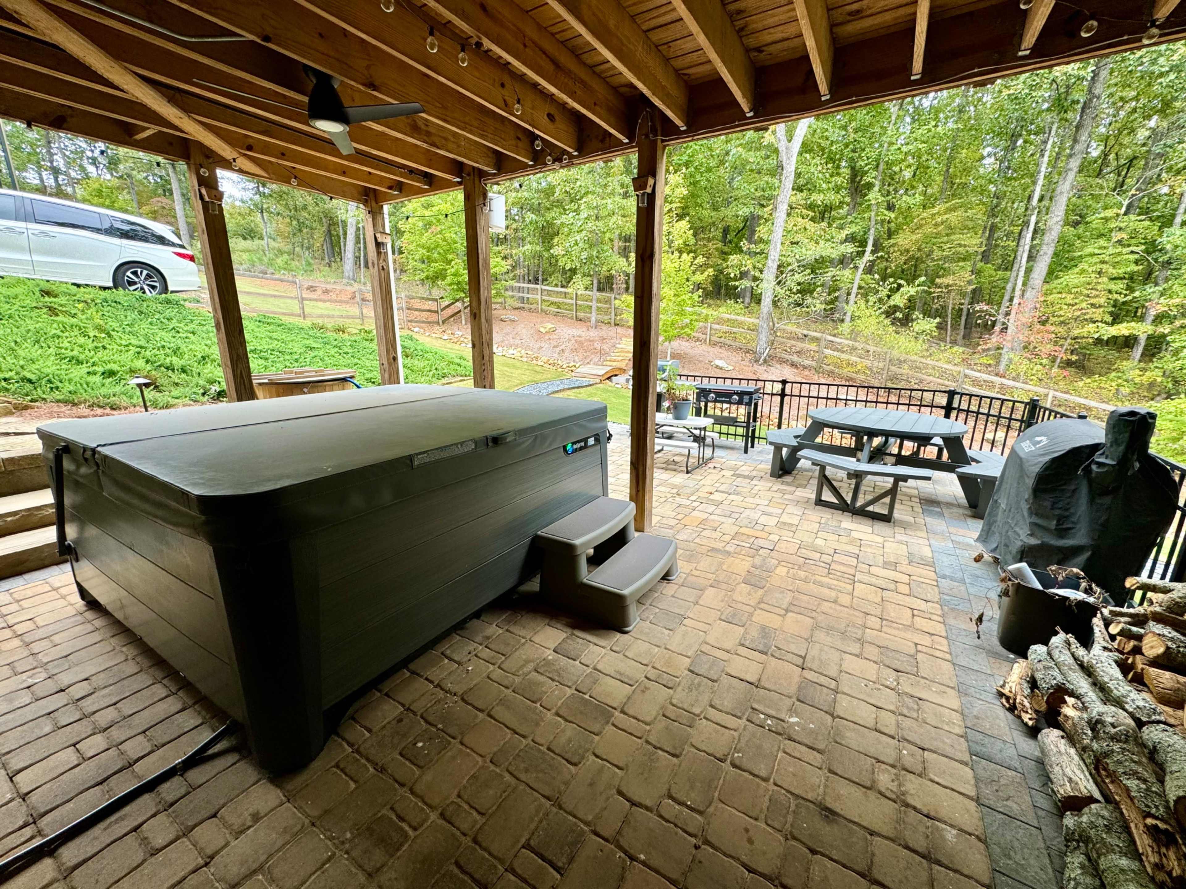 A covered patio area featuring a hot tub, a picnic table, a grill, and a stack of firewood, with a wooded backyard visible in the background.