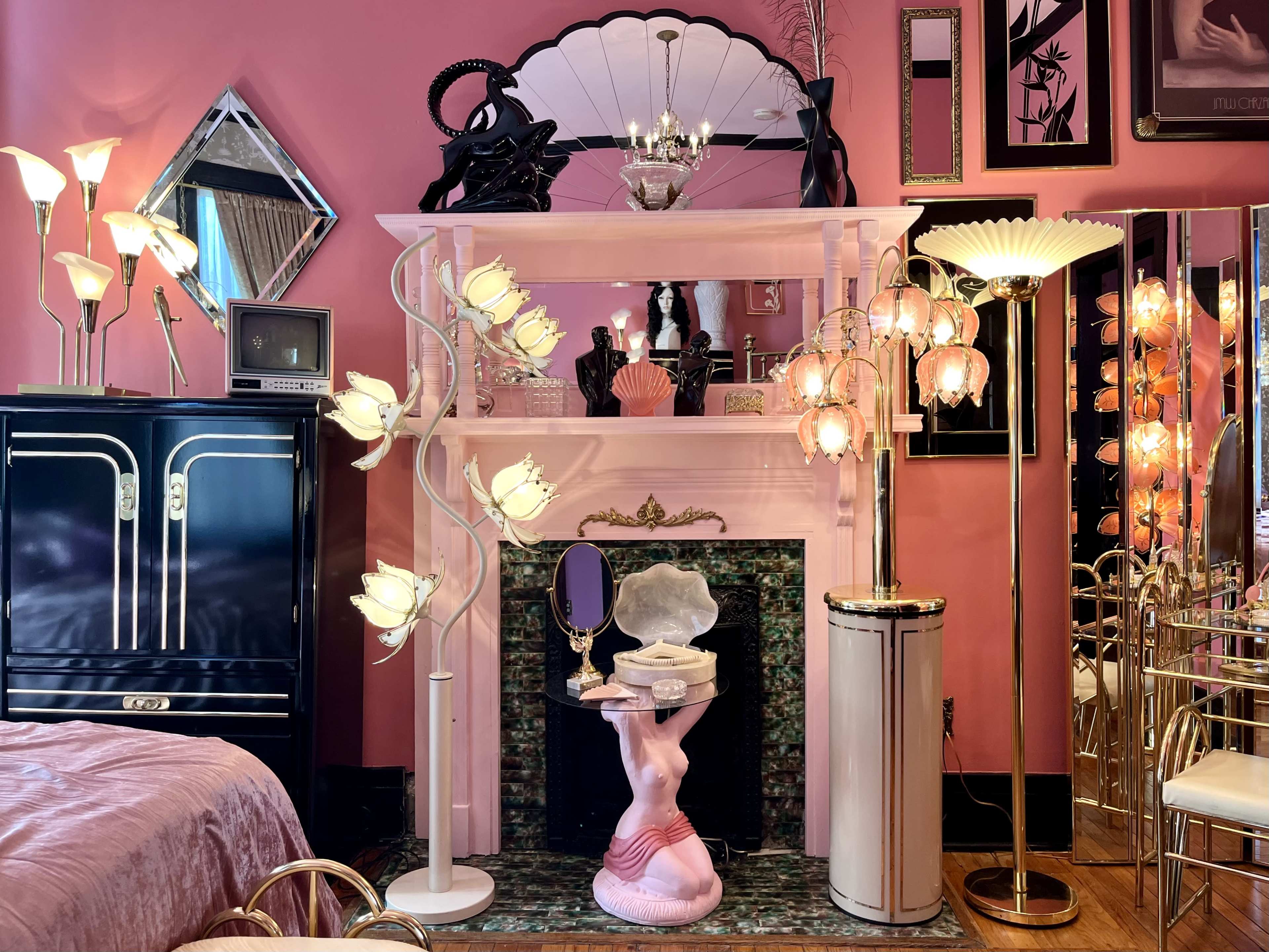 The image shows a decorative room with a pink wall, featuring a fireplace adorned with various lamps, a mirror, and a pink sculpture of a female figure.
