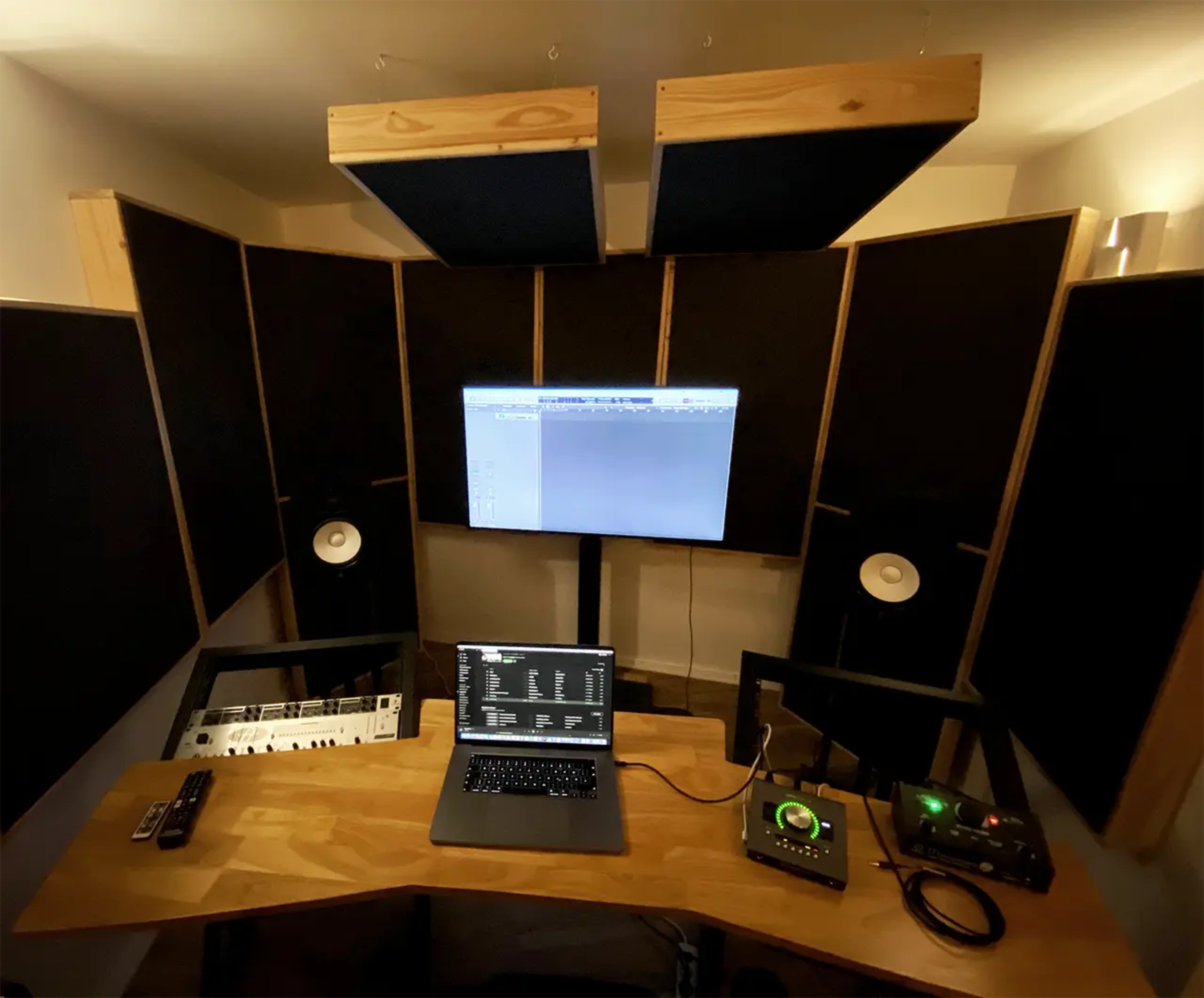 A music production studio is set up with a computer, audio interface, MIDI keyboard, and speakers arranged on a wooden desk surrounded by acoustic panels.