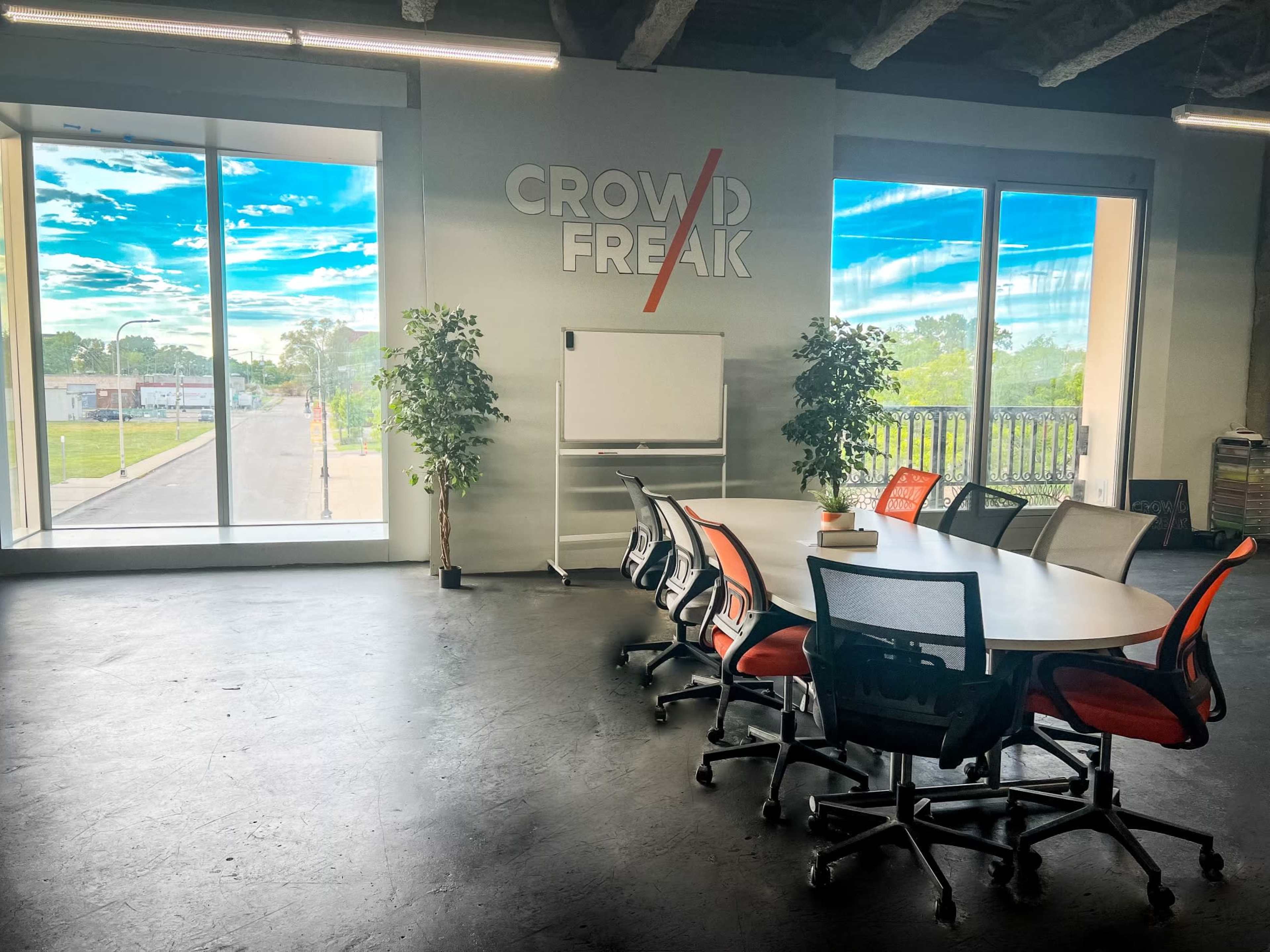 A conference room features a large round table surrounded by chairs, with floor-to-ceiling windows showing an outdoor view and a wall displaying the text "CROWD FREAK."