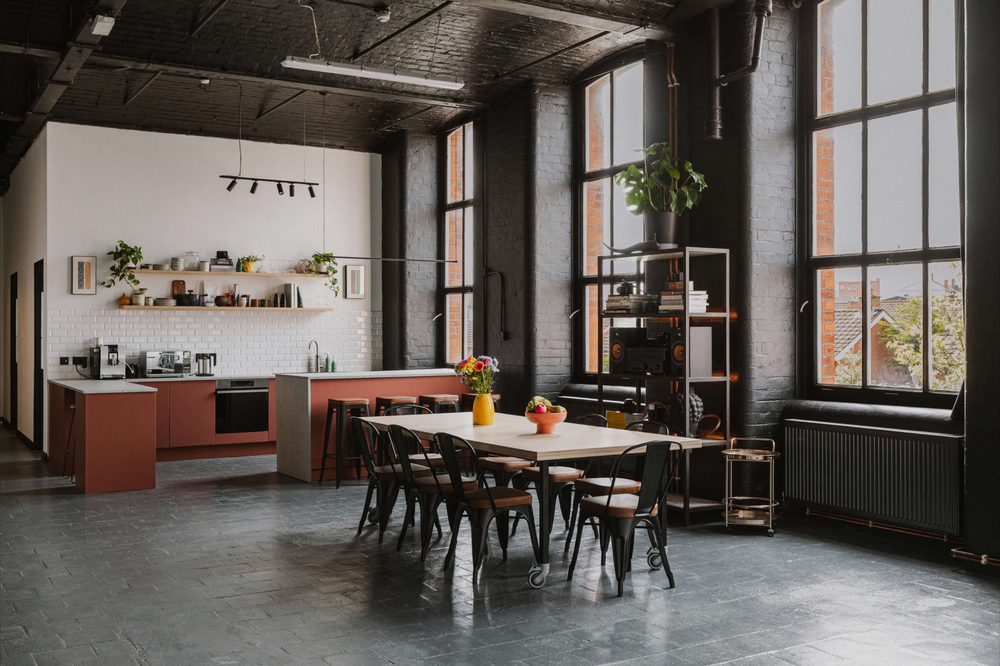 A modern kitchen and dining area features large windows, a long wooden table with black chairs, and a sleek kitchen with a pink and gray color scheme.