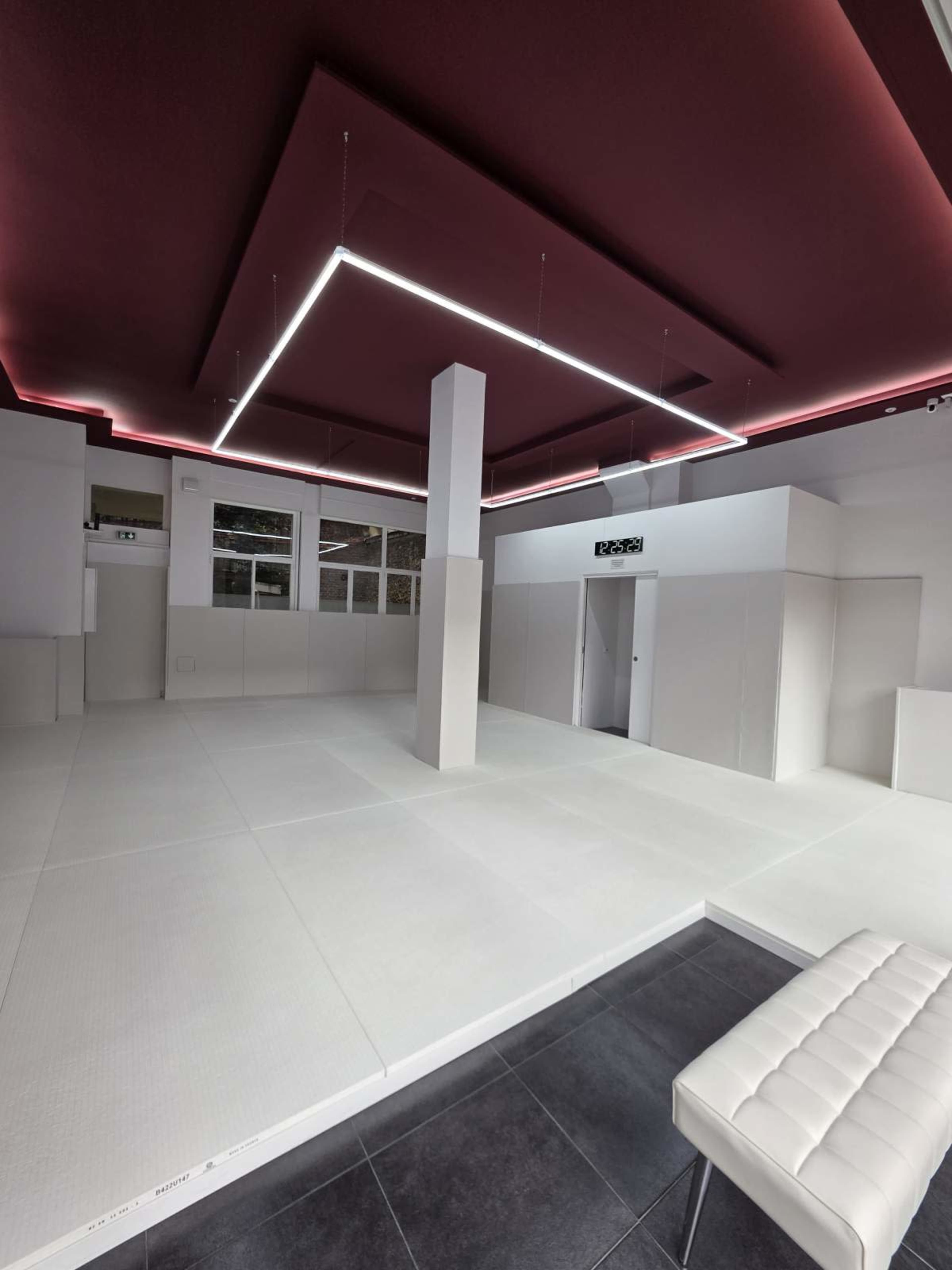 The image depicts a modern, minimalistic interior space with white padded walls, a dark tiled floor, and a ceiling featuring geometric lighting.