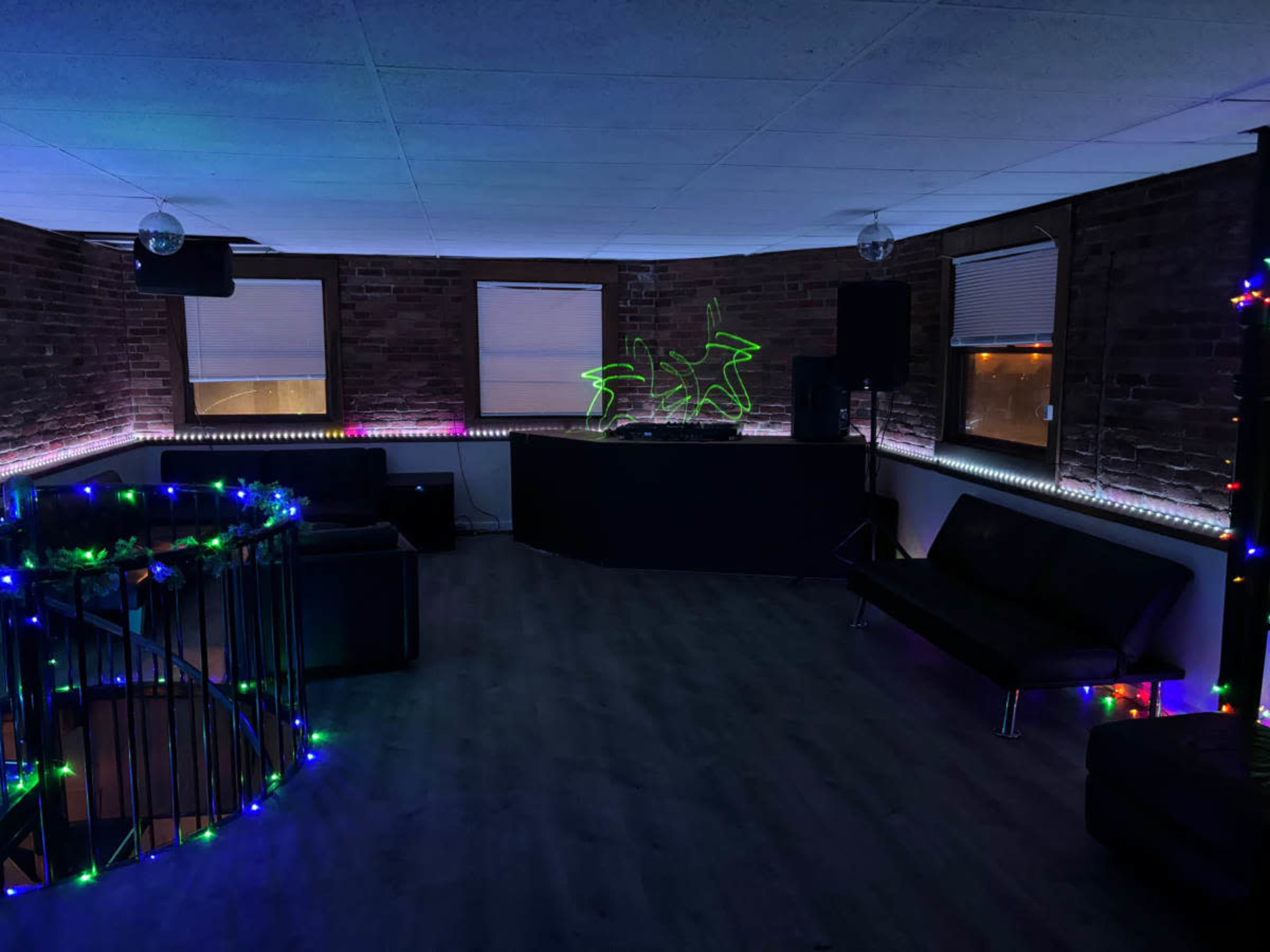 The image shows a dimly lit room with brick walls, featuring a DJ setup, two couches, and colorful lights along the edges of the space.