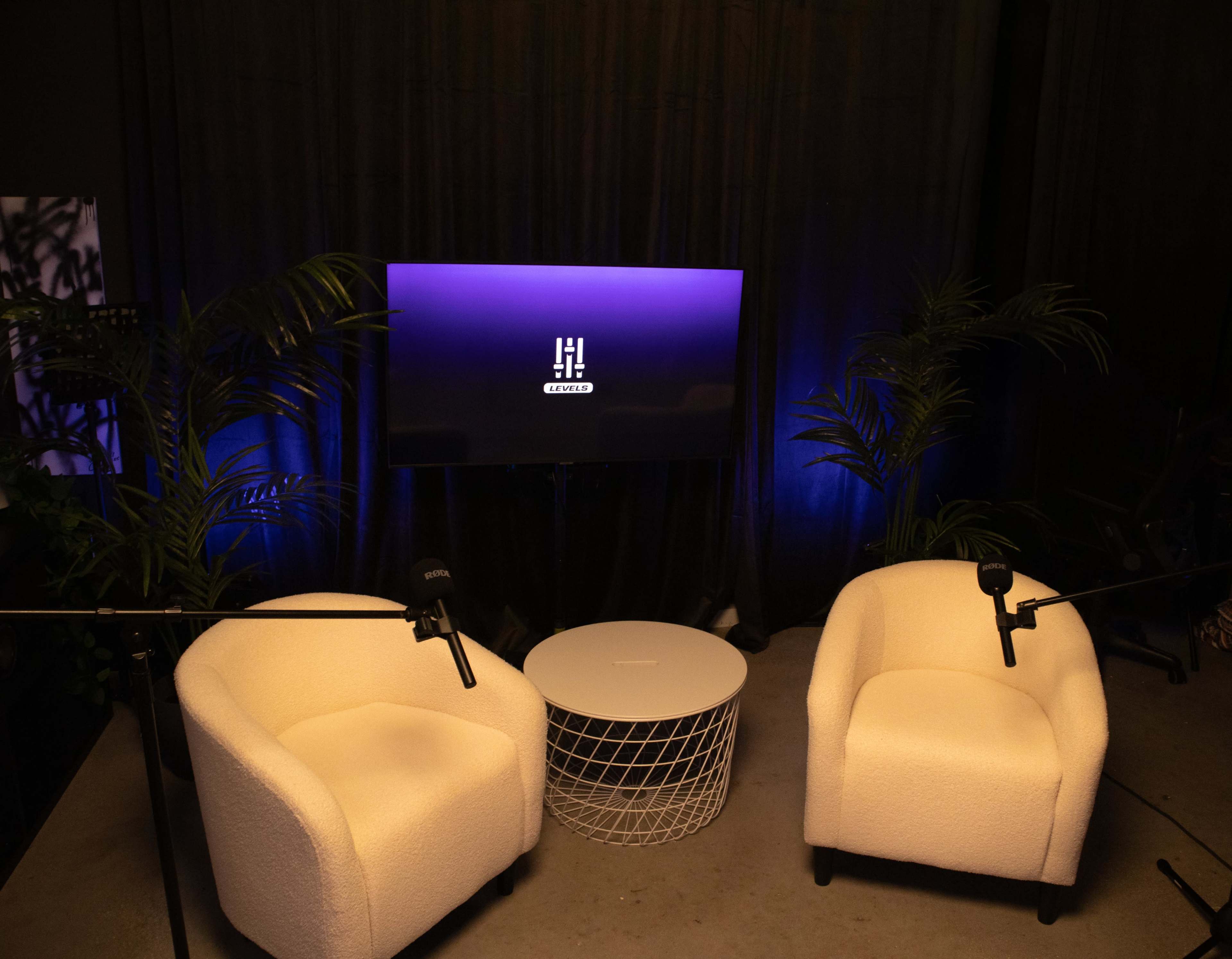 The image shows a podcast or interview setup featuring two white armchairs, a small round table, and a screen displaying a logo, with surrounding plants and dim lighting.