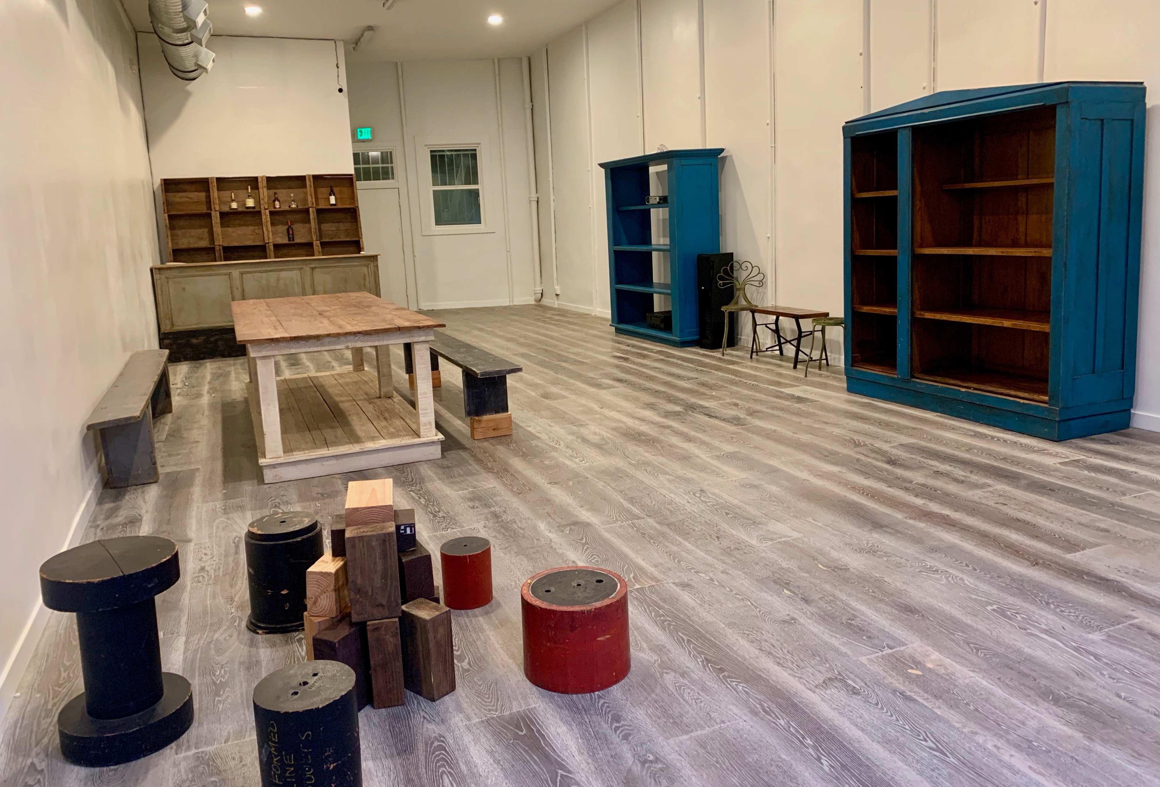 An empty room with wooden flooring, two blue bookcases, a wooden table, various cylindrical stools, and a set of shelves against the wall.