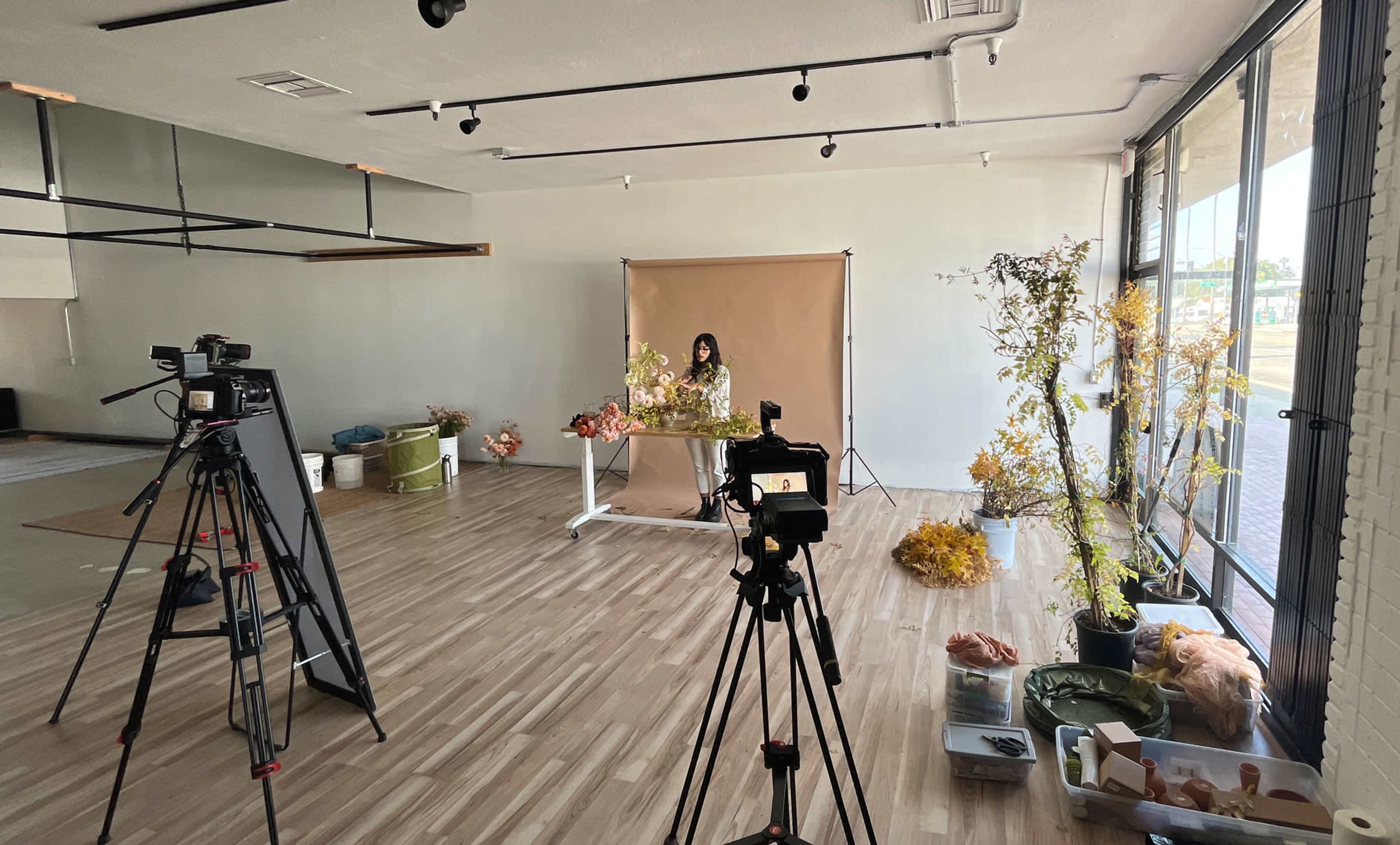 A photographer is set up in a bright studio with multiple cameras focused on a woman arranging dried flowers against a backdrop.
