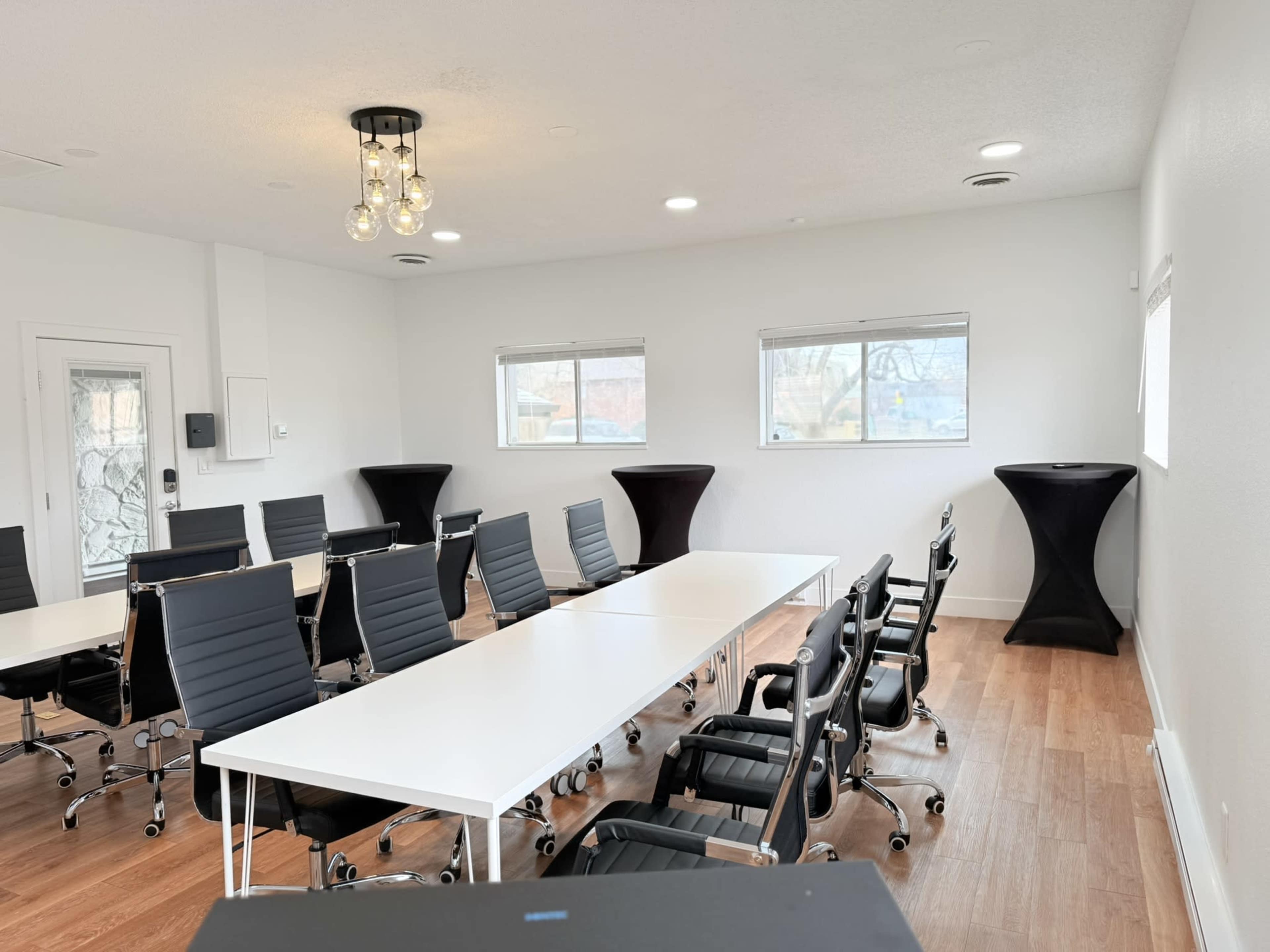 The image shows a modern conference room with a long white table surrounded by black chairs, along with two high tables at the sides.