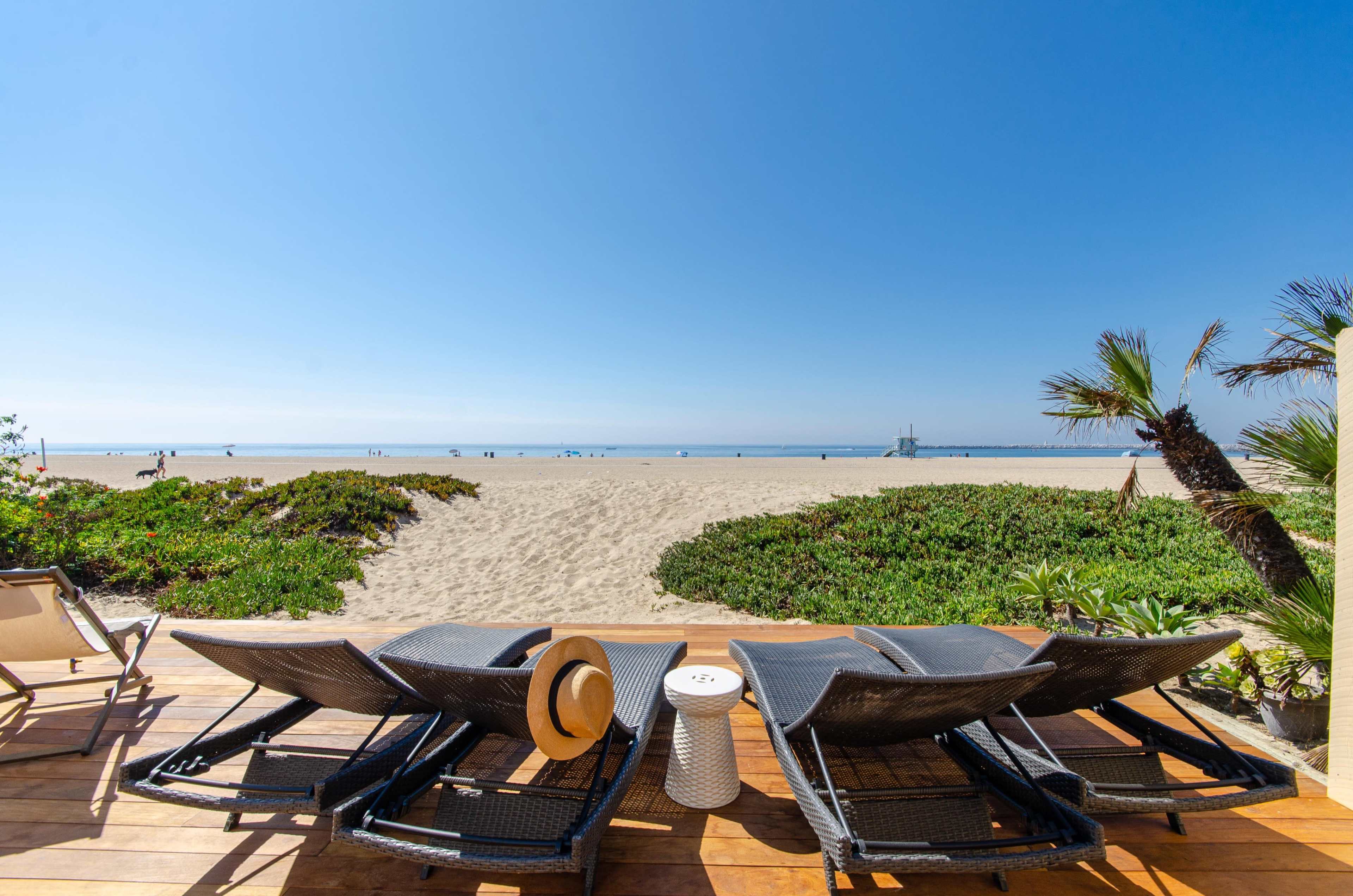 A row of lounge chairs is positioned on a wooden deck overlooking a sandy beach and calm ocean under a clear blue sky.