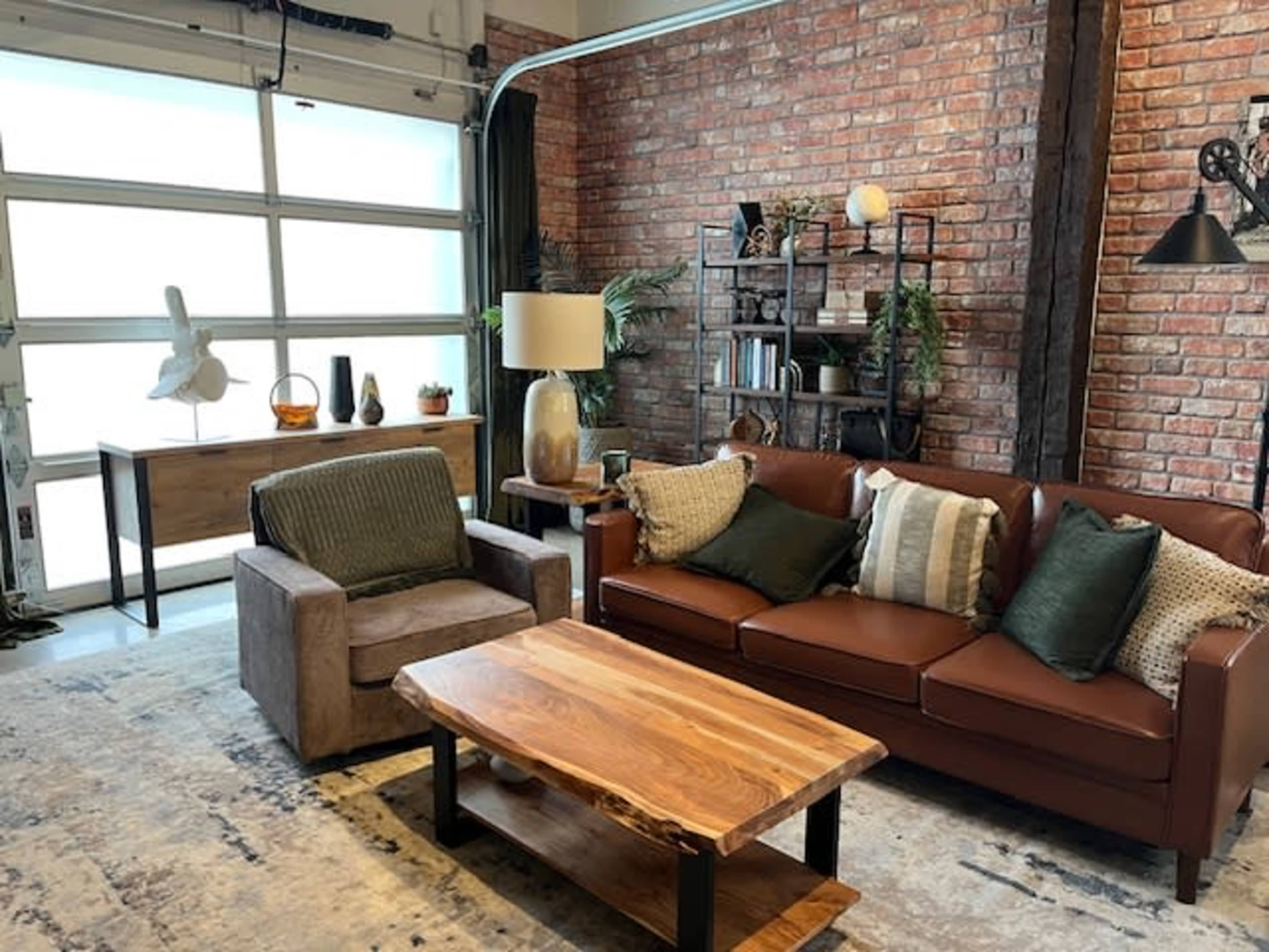 The image shows a living room with a brown leather sofa, an armchair, a wooden coffee table, and a windowed garage door, set against a backdrop of exposed brick walls and modern decor.