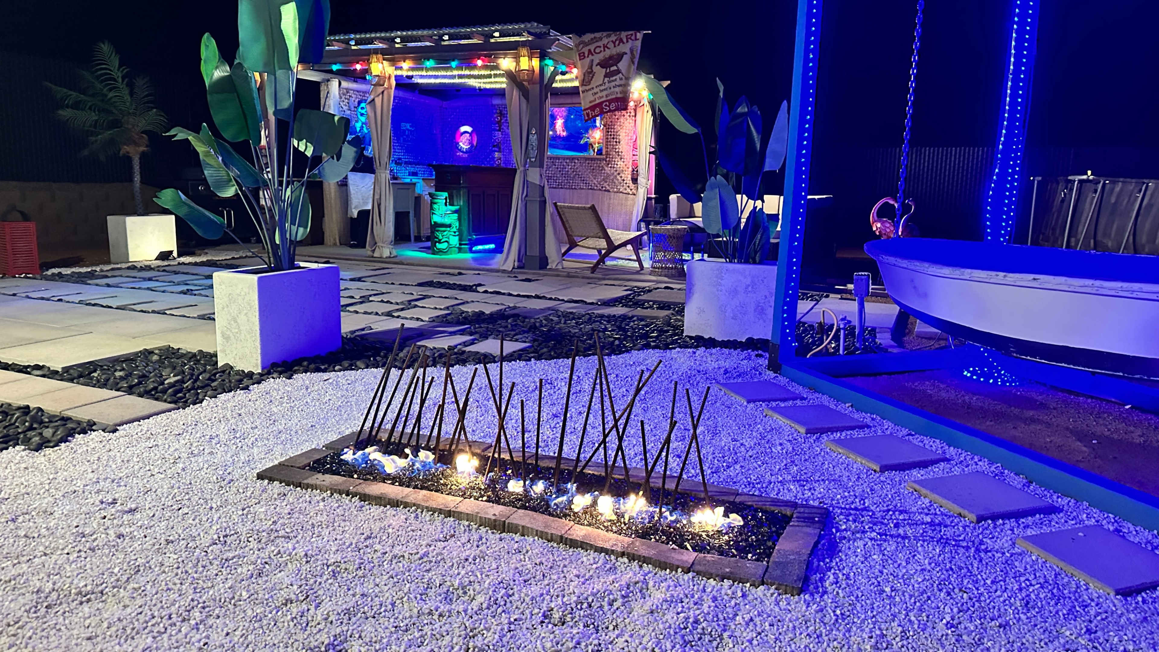 The scene features a well-lit outdoor lounge area with colorful lighting, a thatched-roof bar, and a fire pit surrounded by gravel and stone pathways.