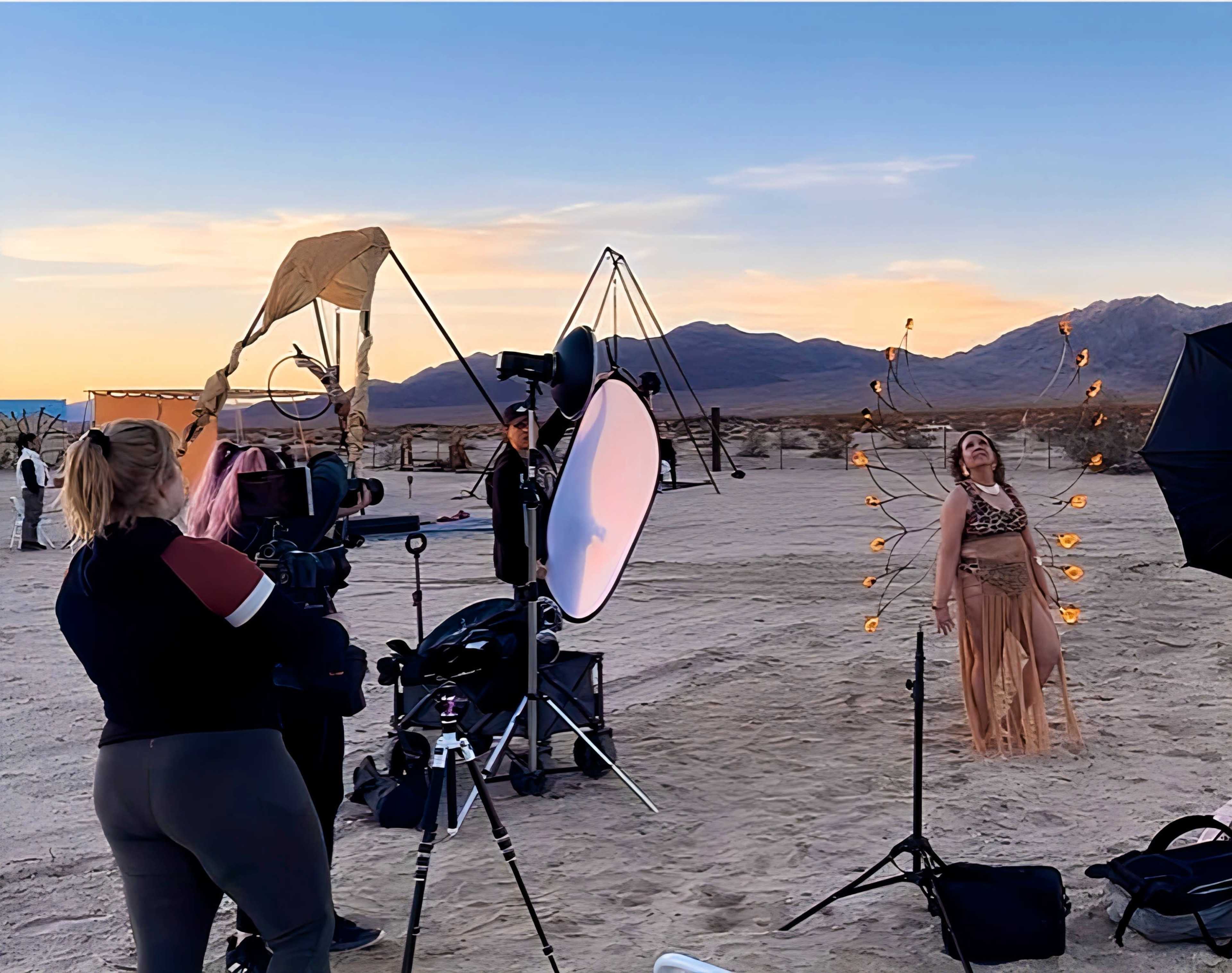 A photoshoot is taking place in a desert landscape, featuring a model in a flowing outfit surrounded by studio lights and equipment at sunset.
