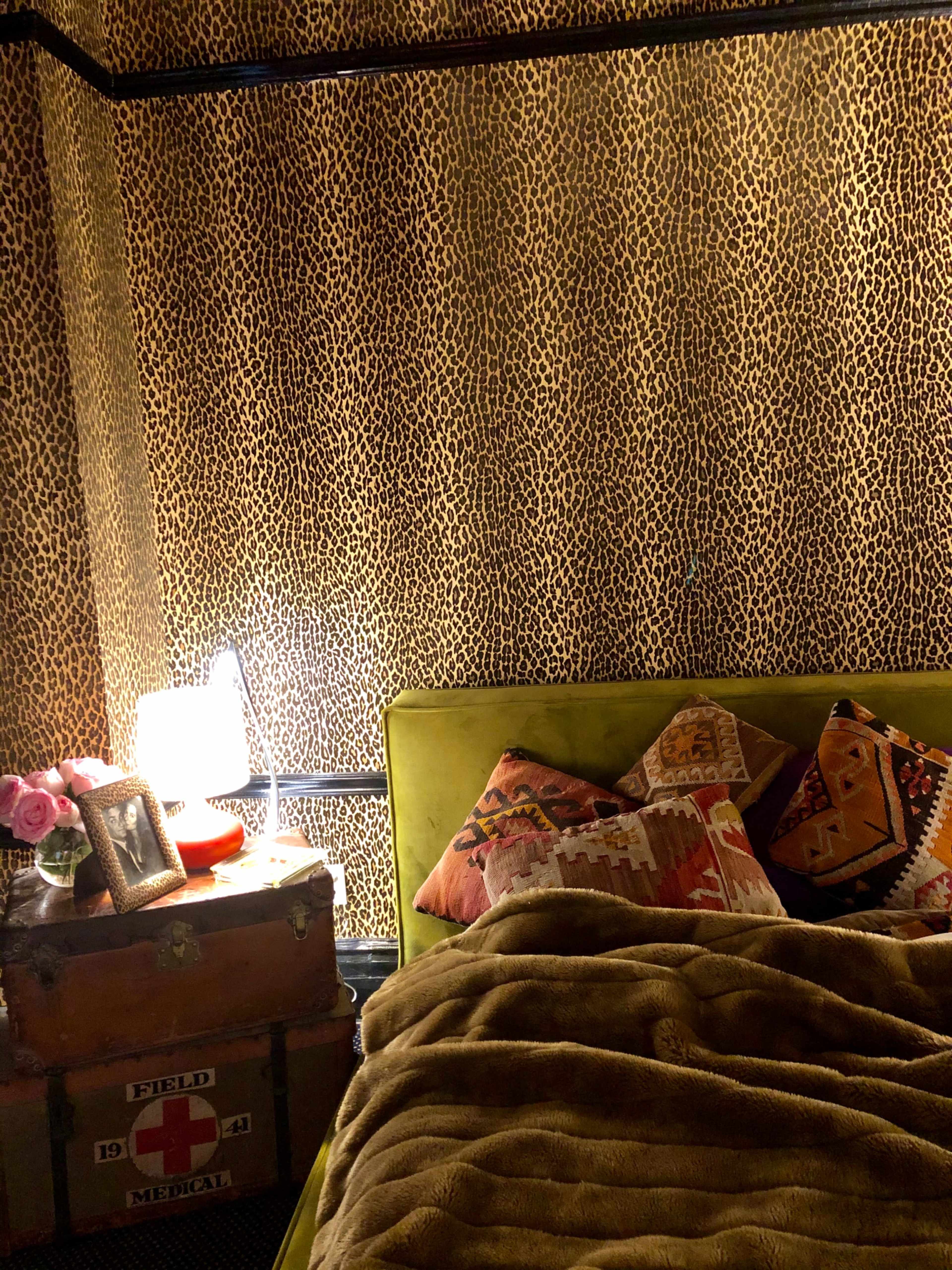 A cozy bedroom with a leopard-print wall, a yellow bed frame, decorative pillows, a bedside lamp, and a vintage trunk.