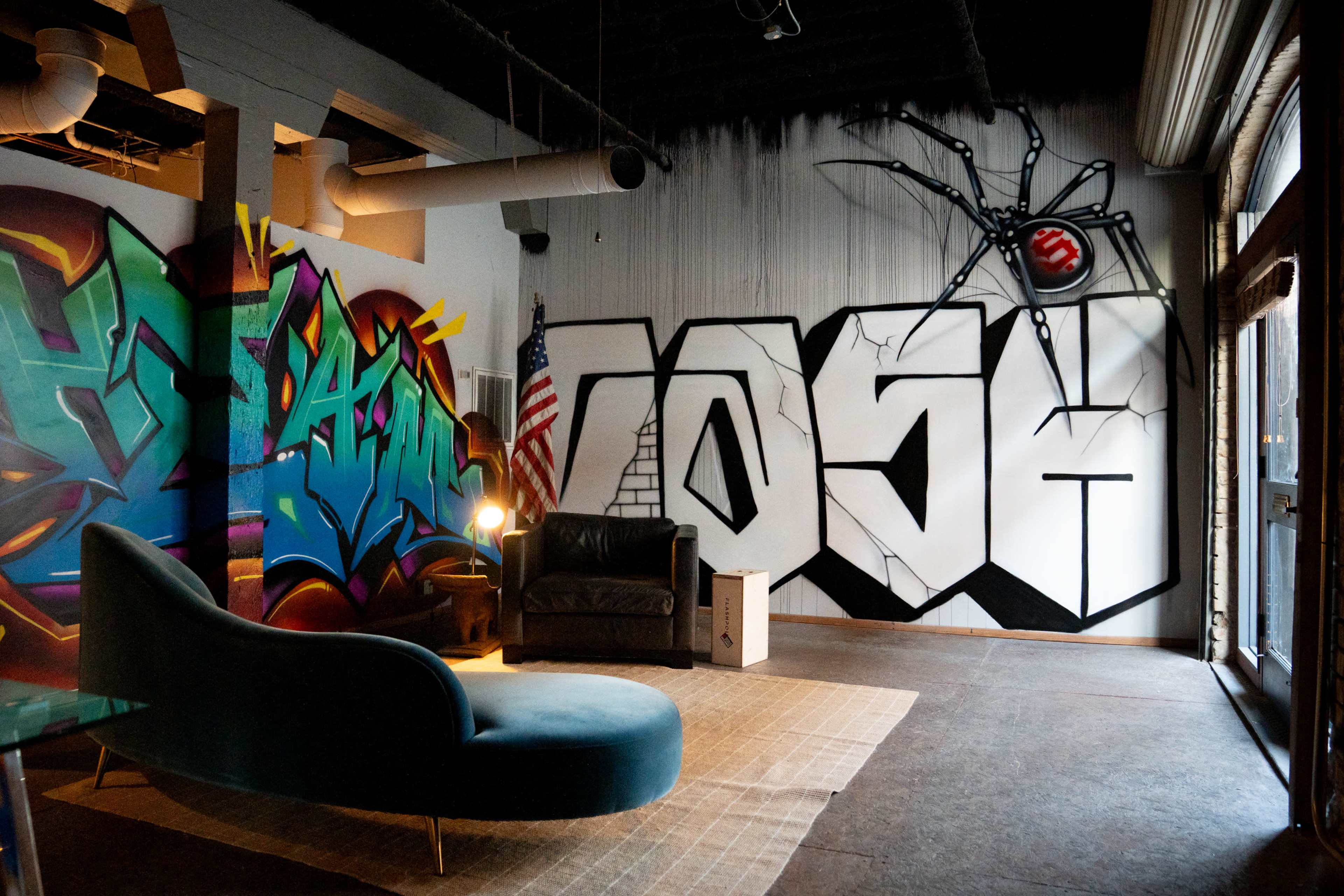 The scene features a modern interior with colorful graffiti on the walls, a dark sofa, a stylish blue couch, and a spider mural near an American flag.