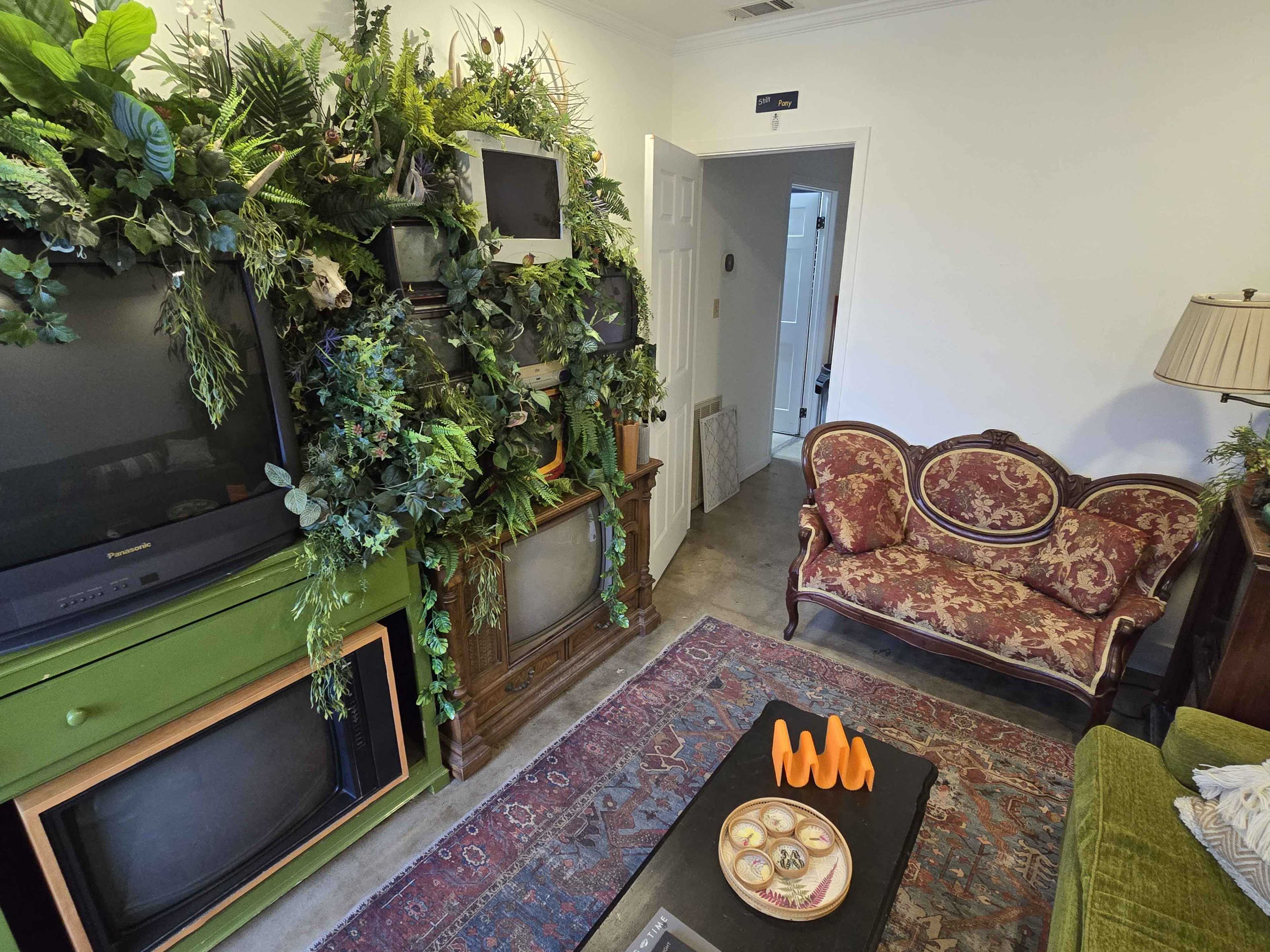 The scene features a small living room with two vintage televisions surrounded by greenery, a patterned sofa, and a coffee table displaying orange decorative items.