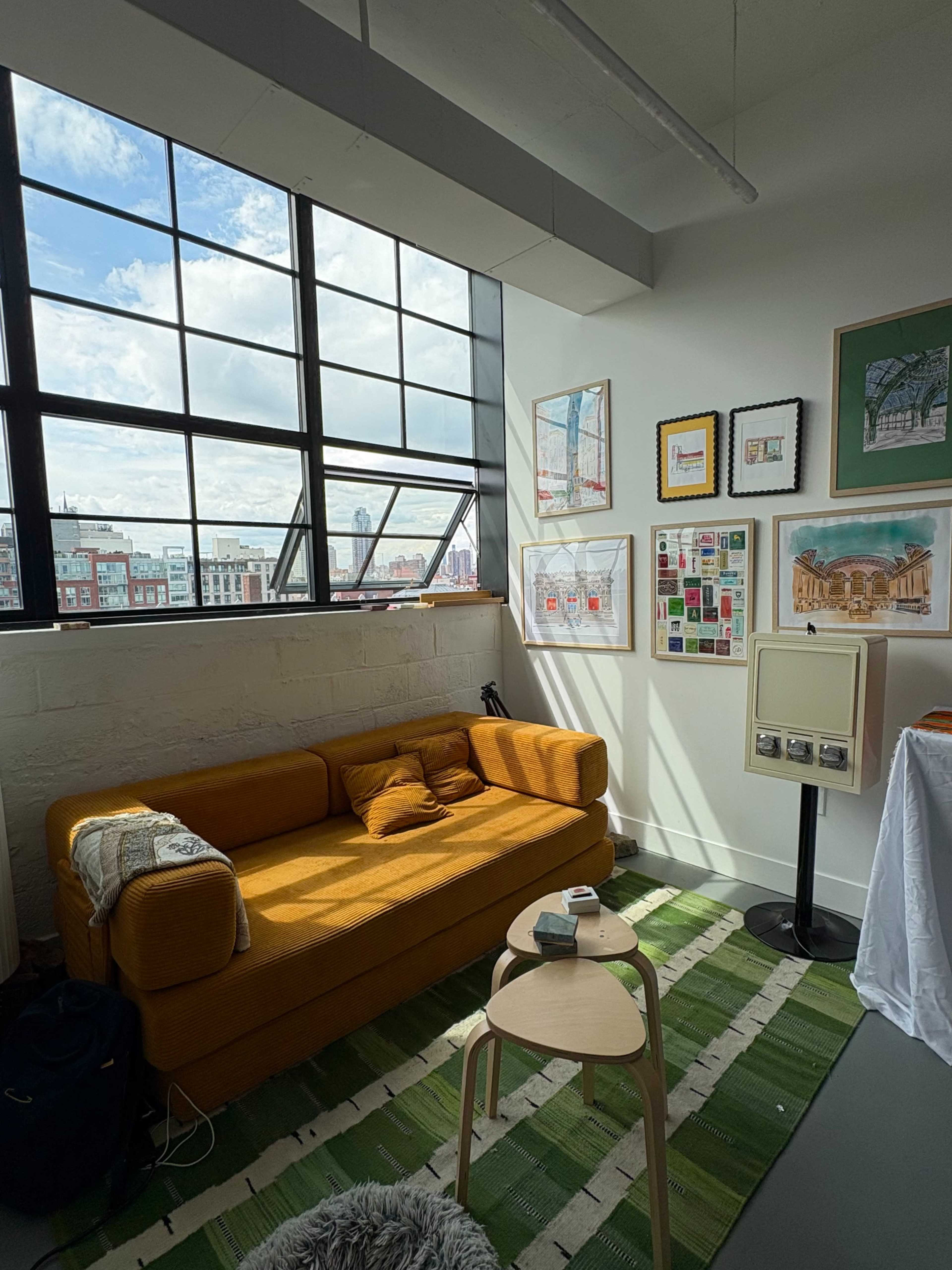 The image shows a bright room featuring a large window, an orange sofa, a wooden side table, and several framed artworks on the walls.