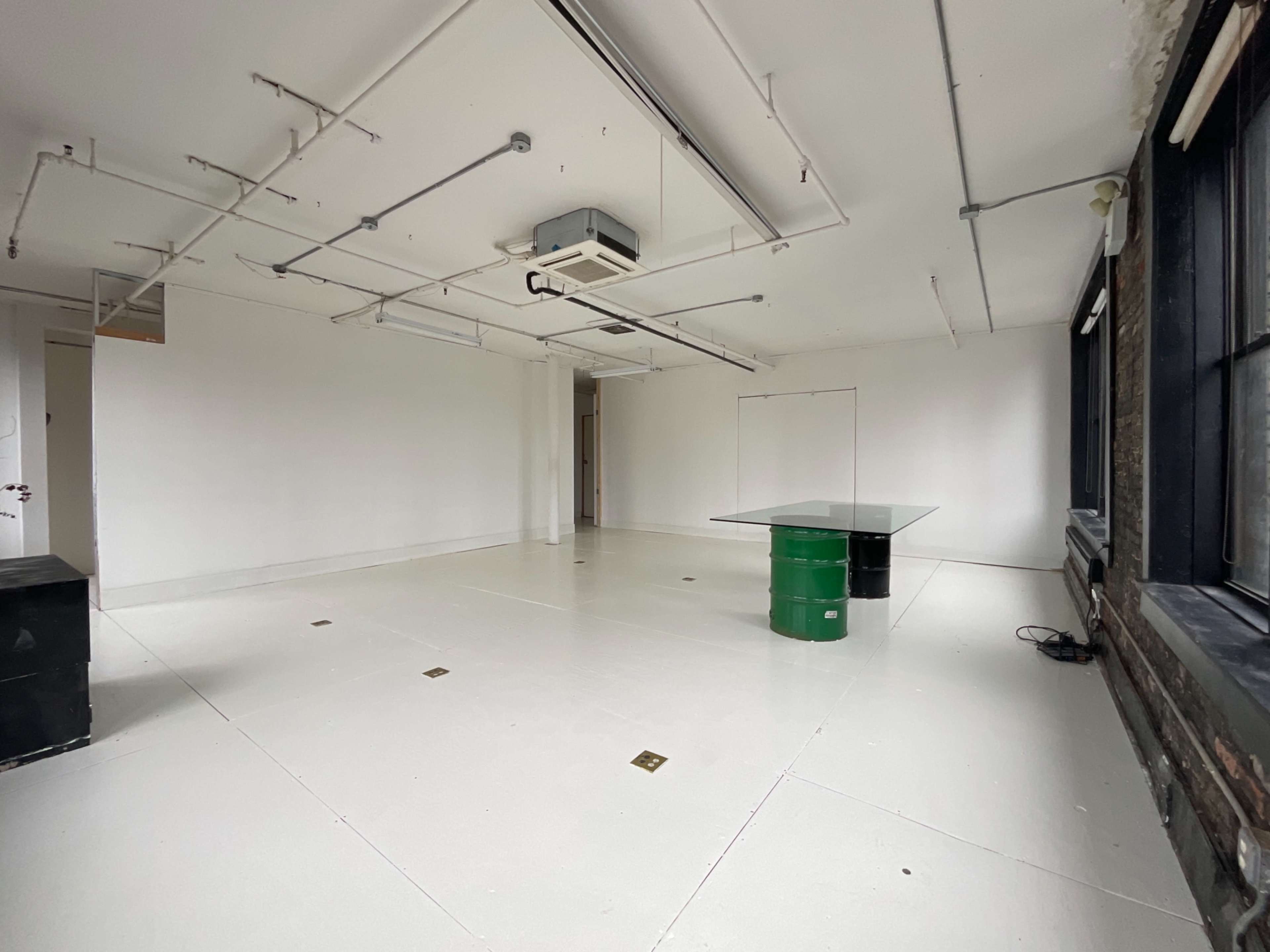 The image shows a spacious room with white walls, a concrete floor, and a glass table positioned on a green cylindrical base.