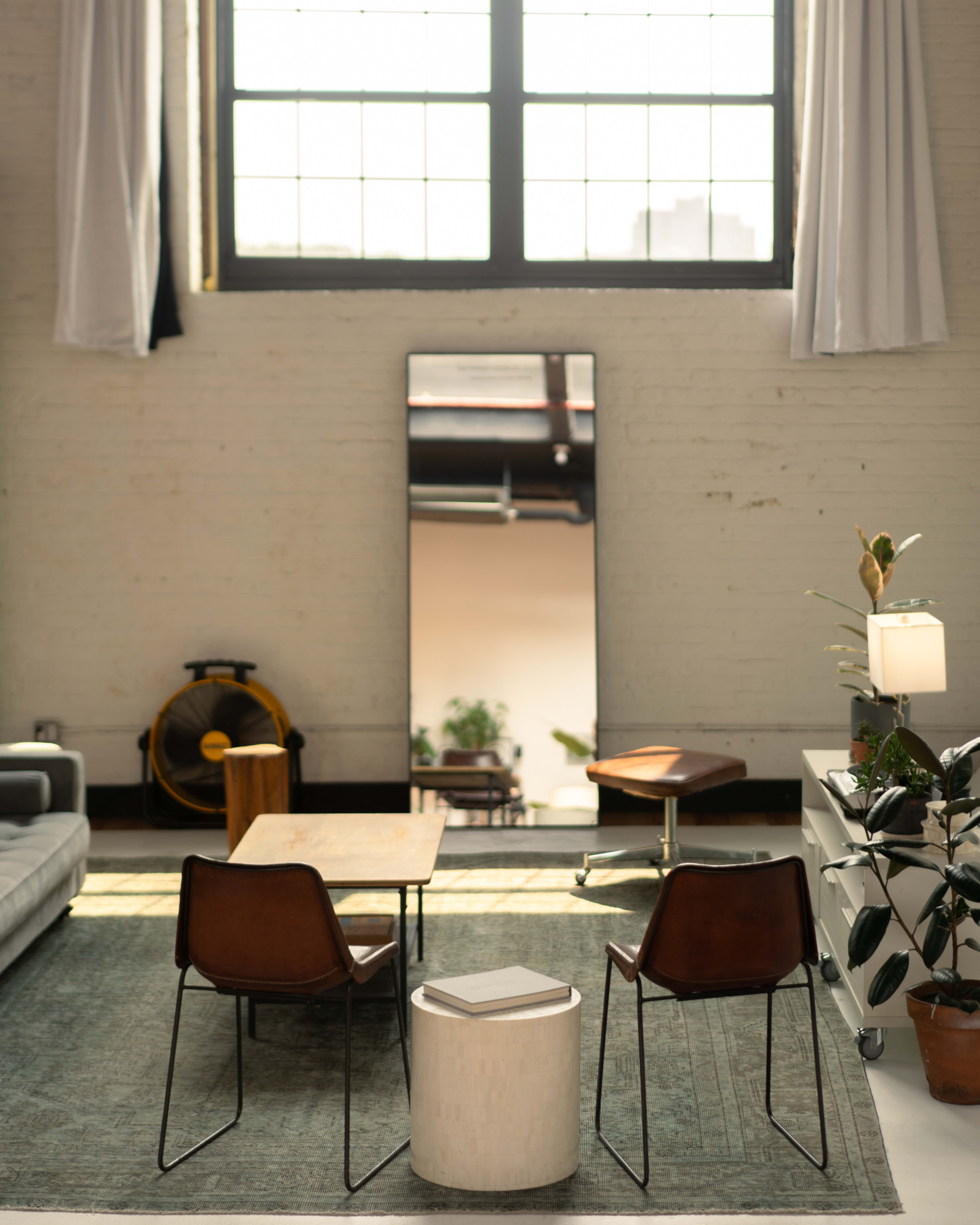 The image showcases a minimalist living space featuring a gray sofa, two brown chairs, a small white table, and a large mirror near a large window.