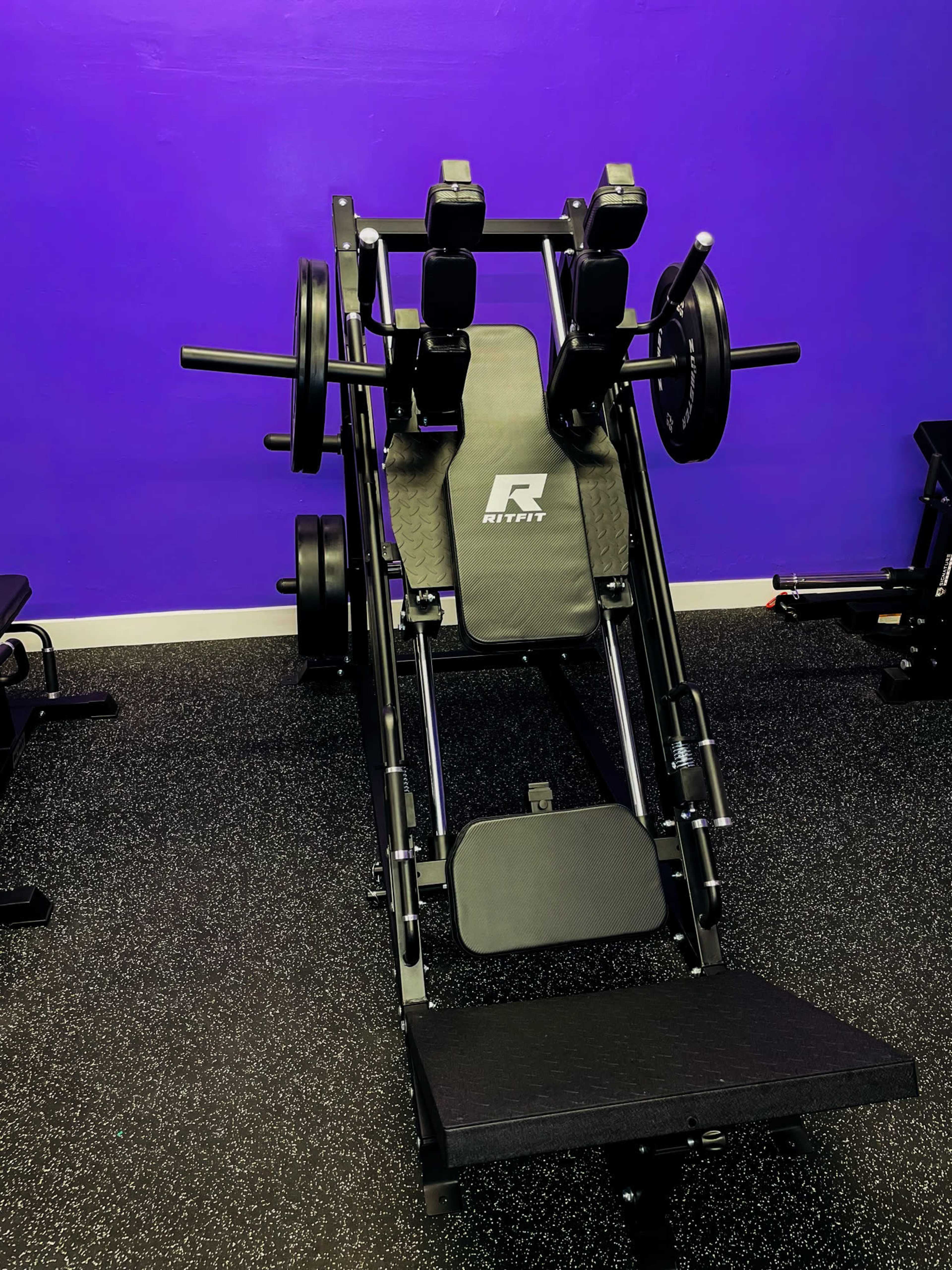 The image shows a gym machine designed for leg exercises, featuring weights, a padded seat, and a purple wall in the background.