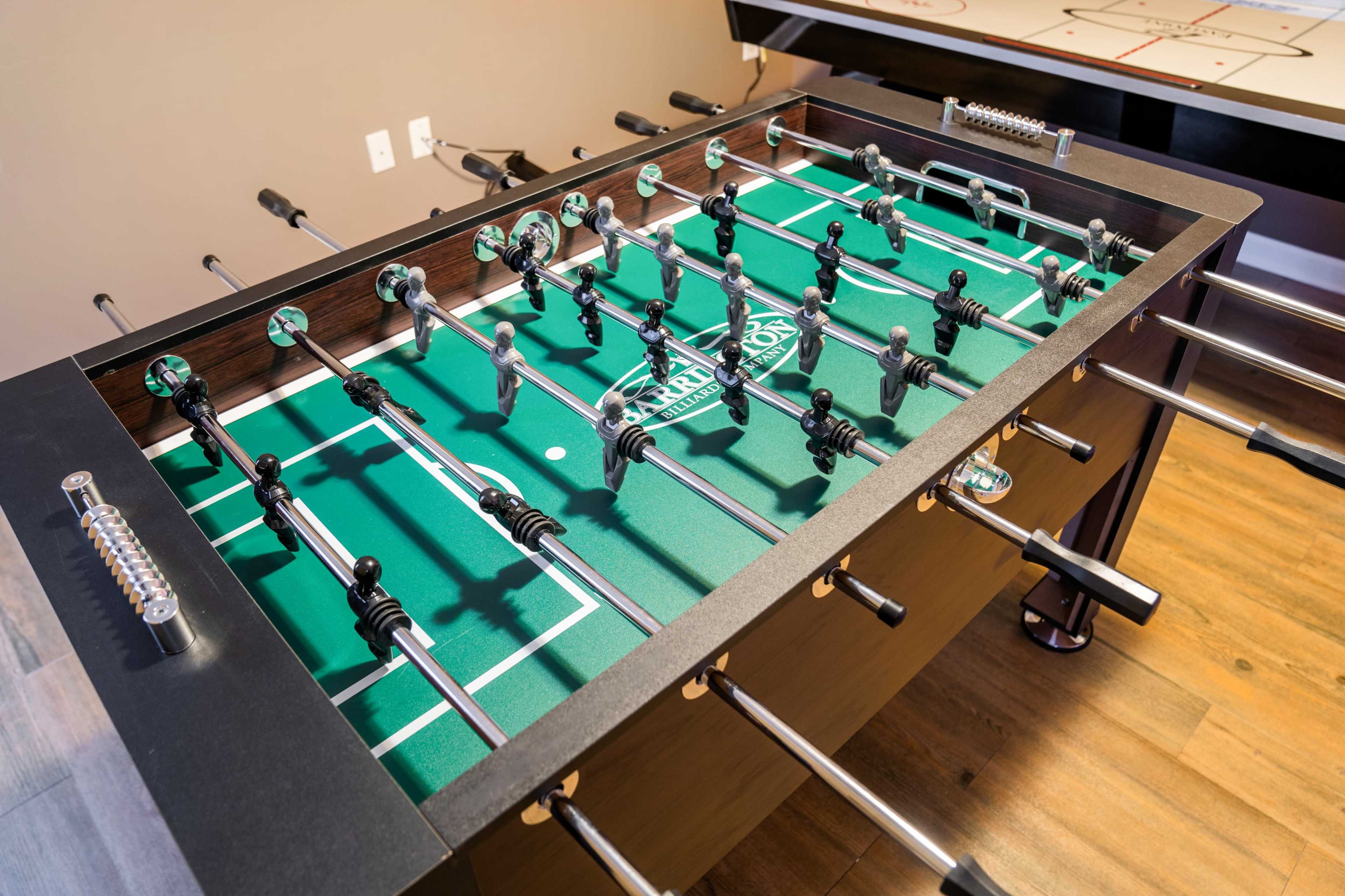 A foosball table with players set up for a game is positioned in a room with wooden flooring and an air hockey table in the background.