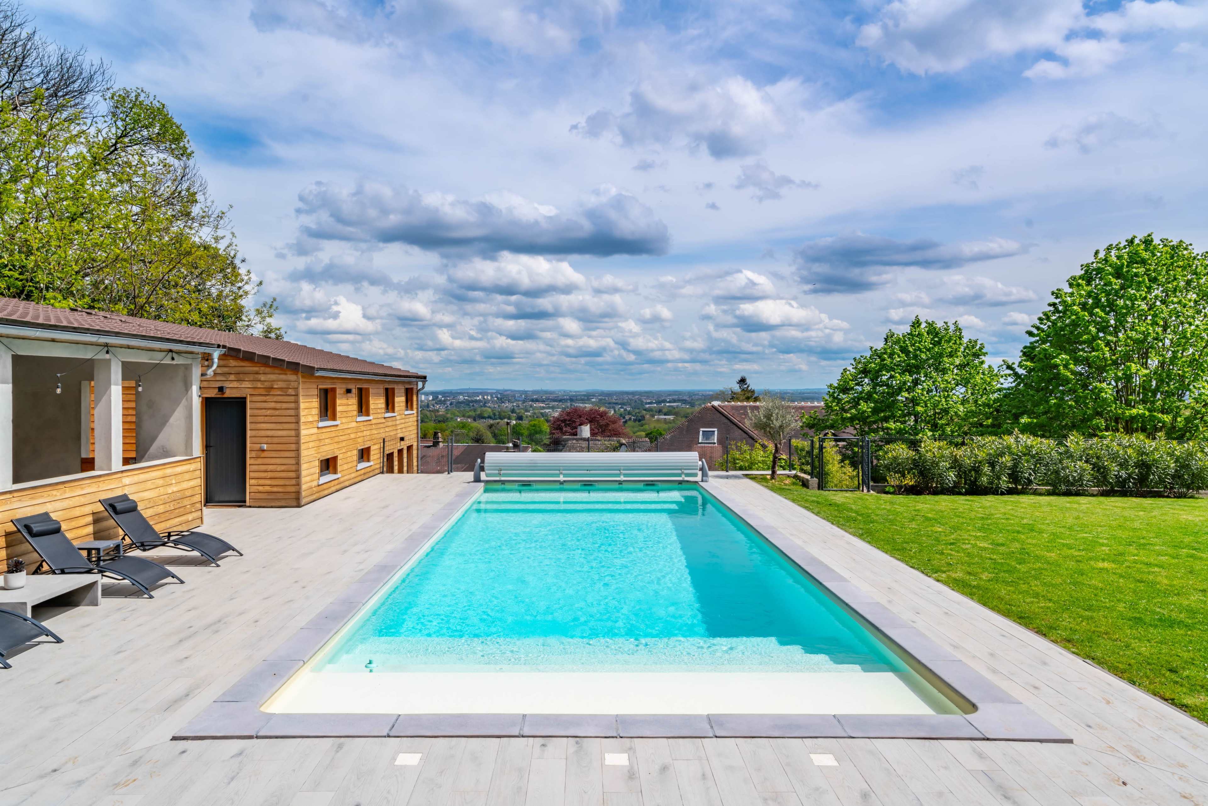 A modern swimming pool with a wooden deck is set against a backdrop of green trees and rolling hills under a partly cloudy sky.