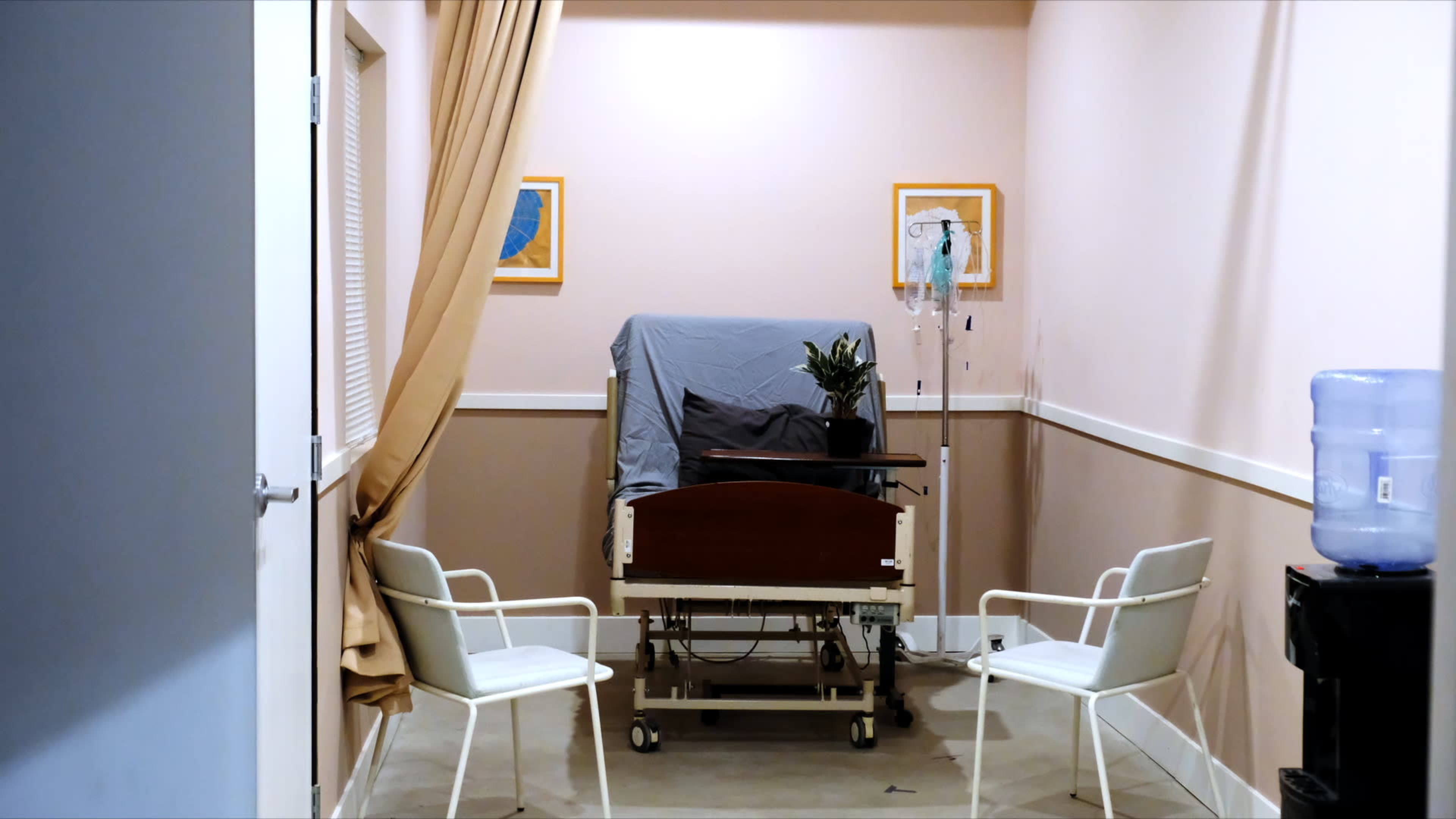 The image shows a small, empty hospital room with a bed covered by a gray blanket, a water cooler in the corner, and two chairs facing the bed.
