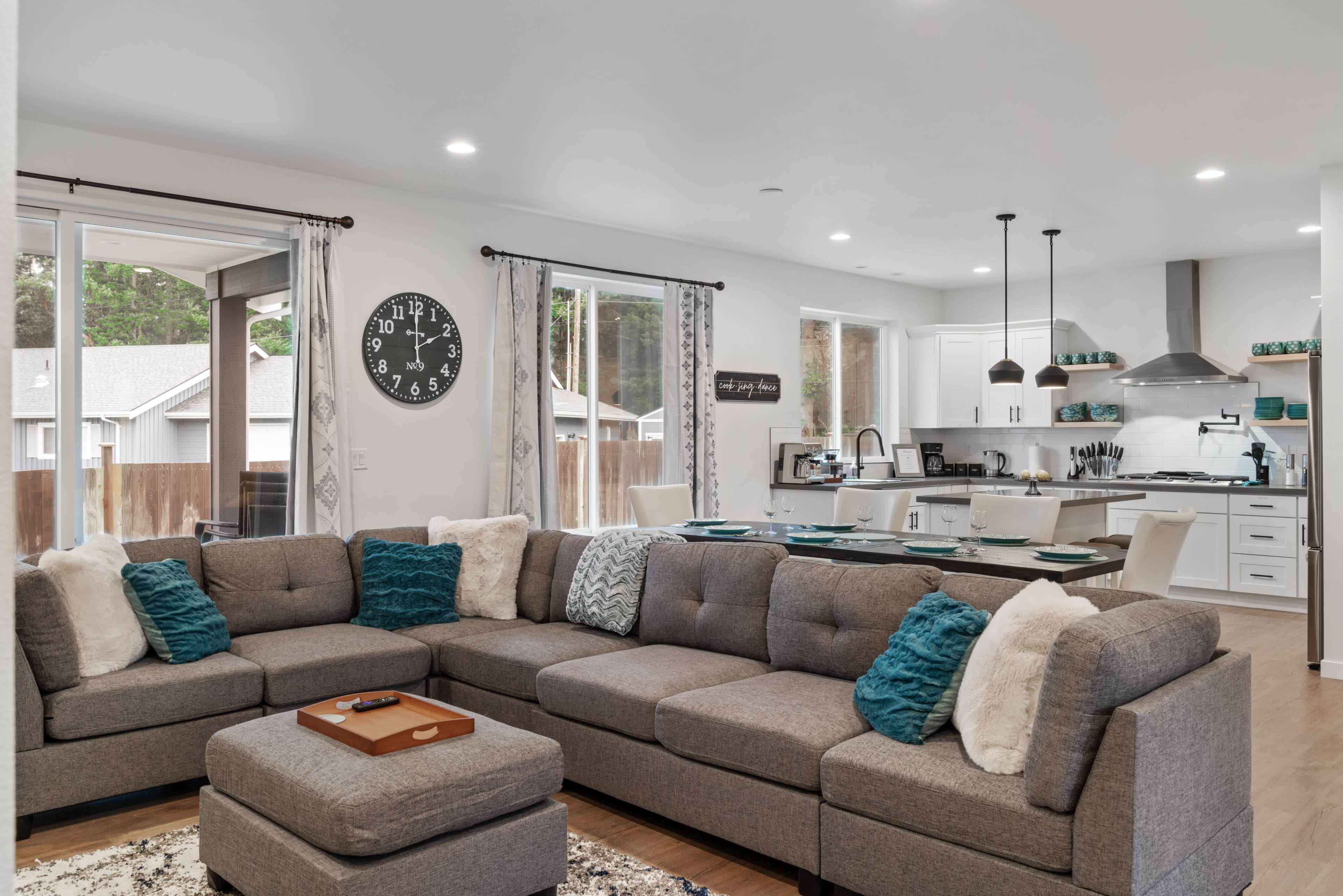 A modern living room features a large gray sectional sofa with decorative pillows, an ottoman, and an open layout connecting to a kitchen area.