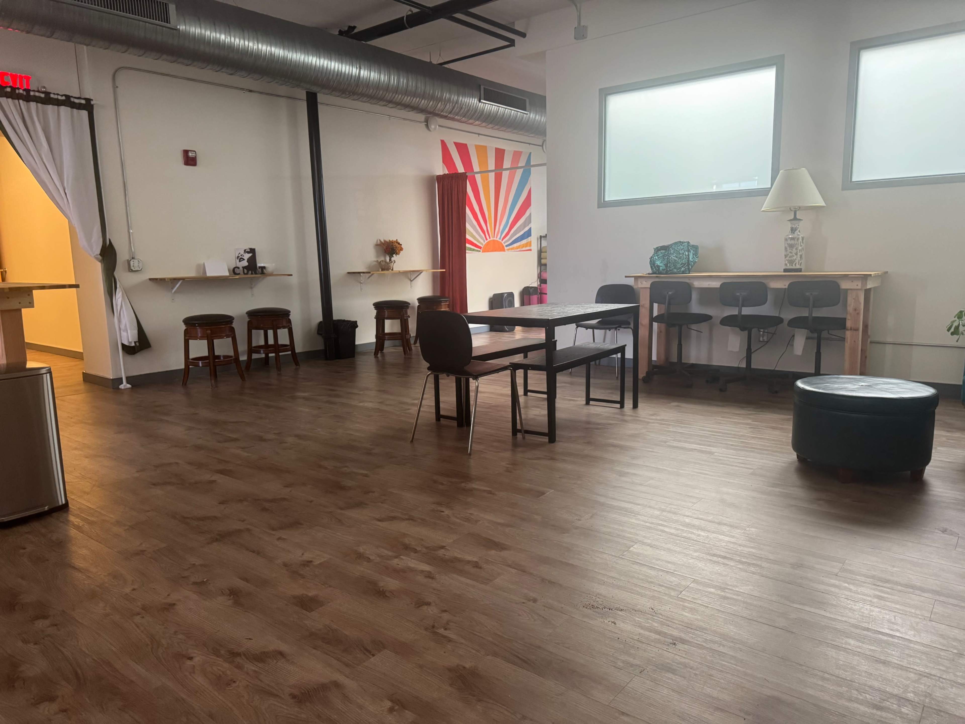 The image shows a spacious room with wooden flooring, featuring a central table, several chairs, and a few small seating areas along the walls.