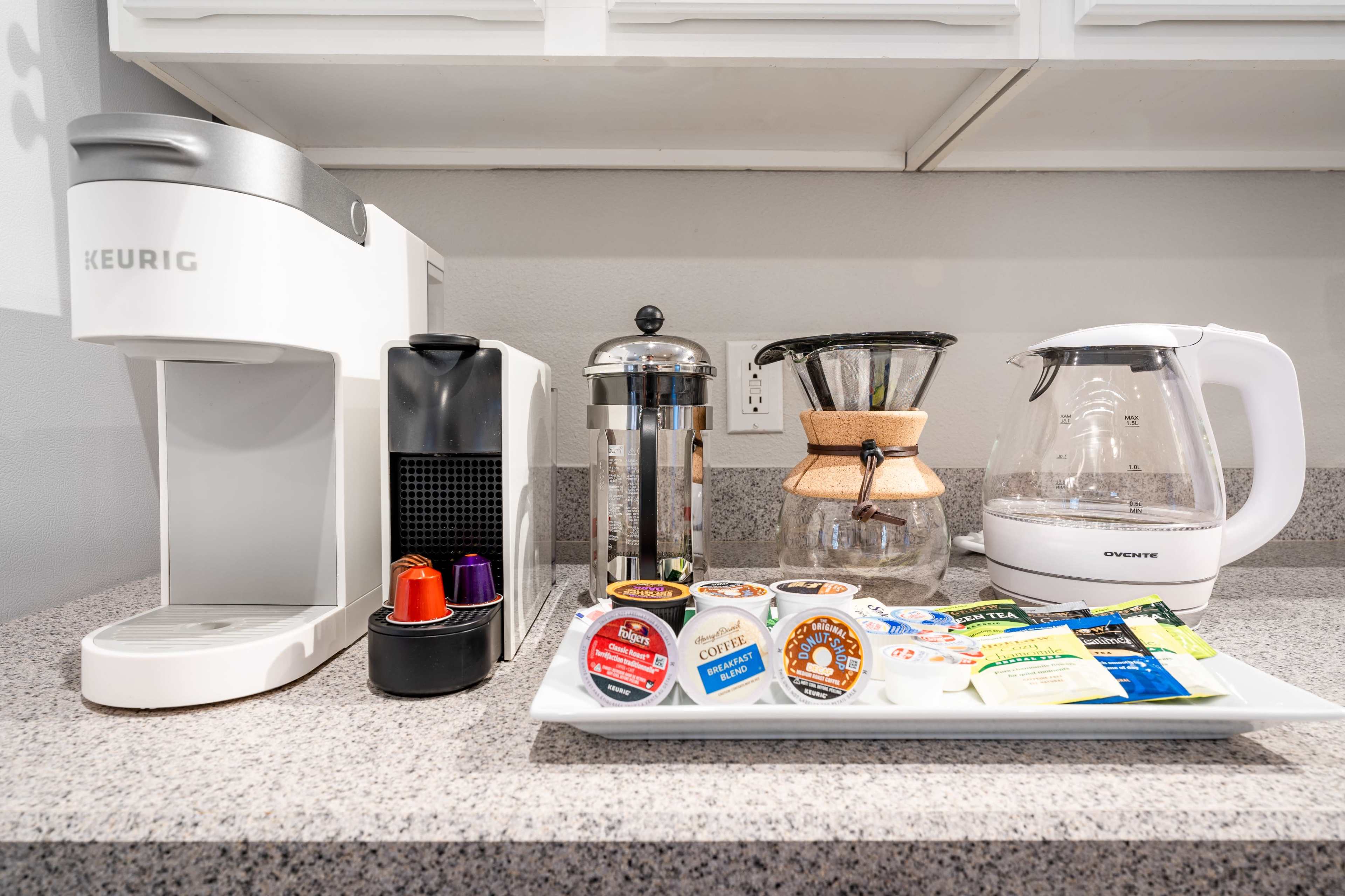 A countertop features a Keurig coffee maker, a French press, an electric kettle, and a tray with various coffee and tea pods.