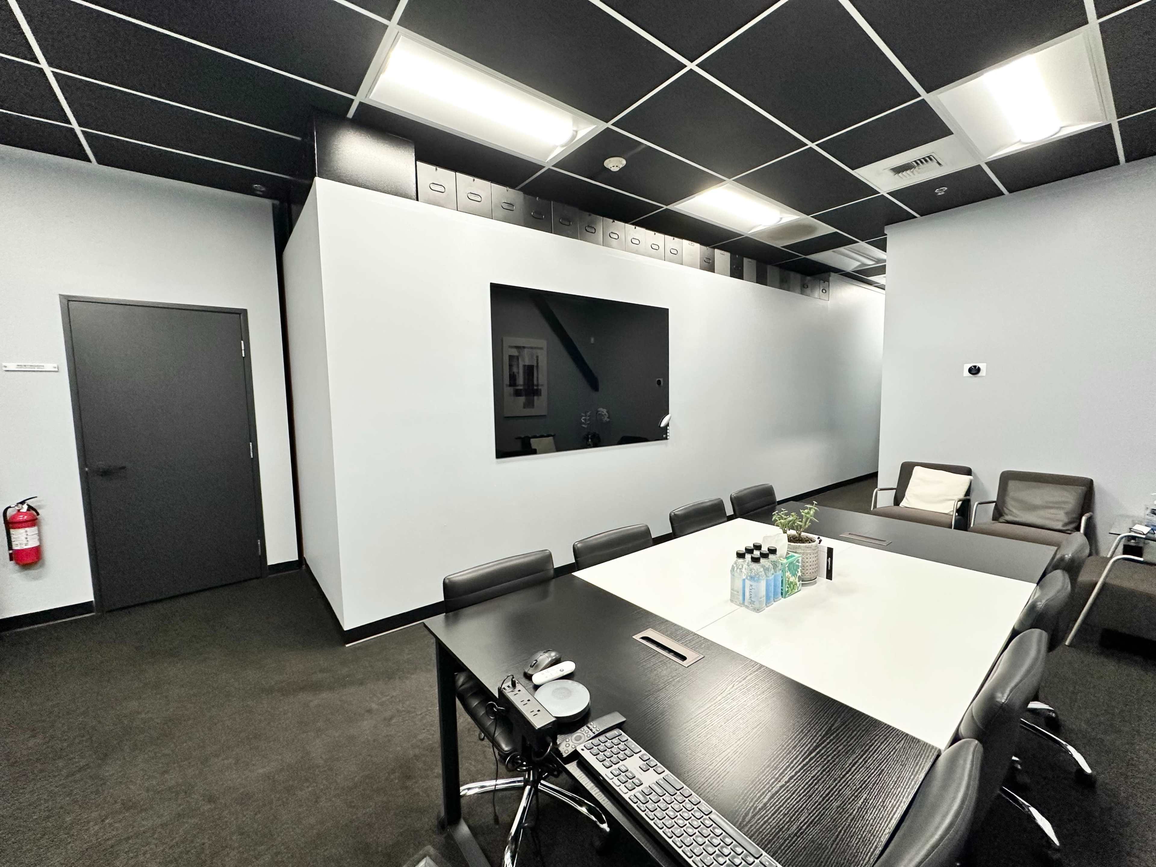 The image shows a modern conference room with a long table, office chairs, a large television mounted on the wall, and a door to the left.