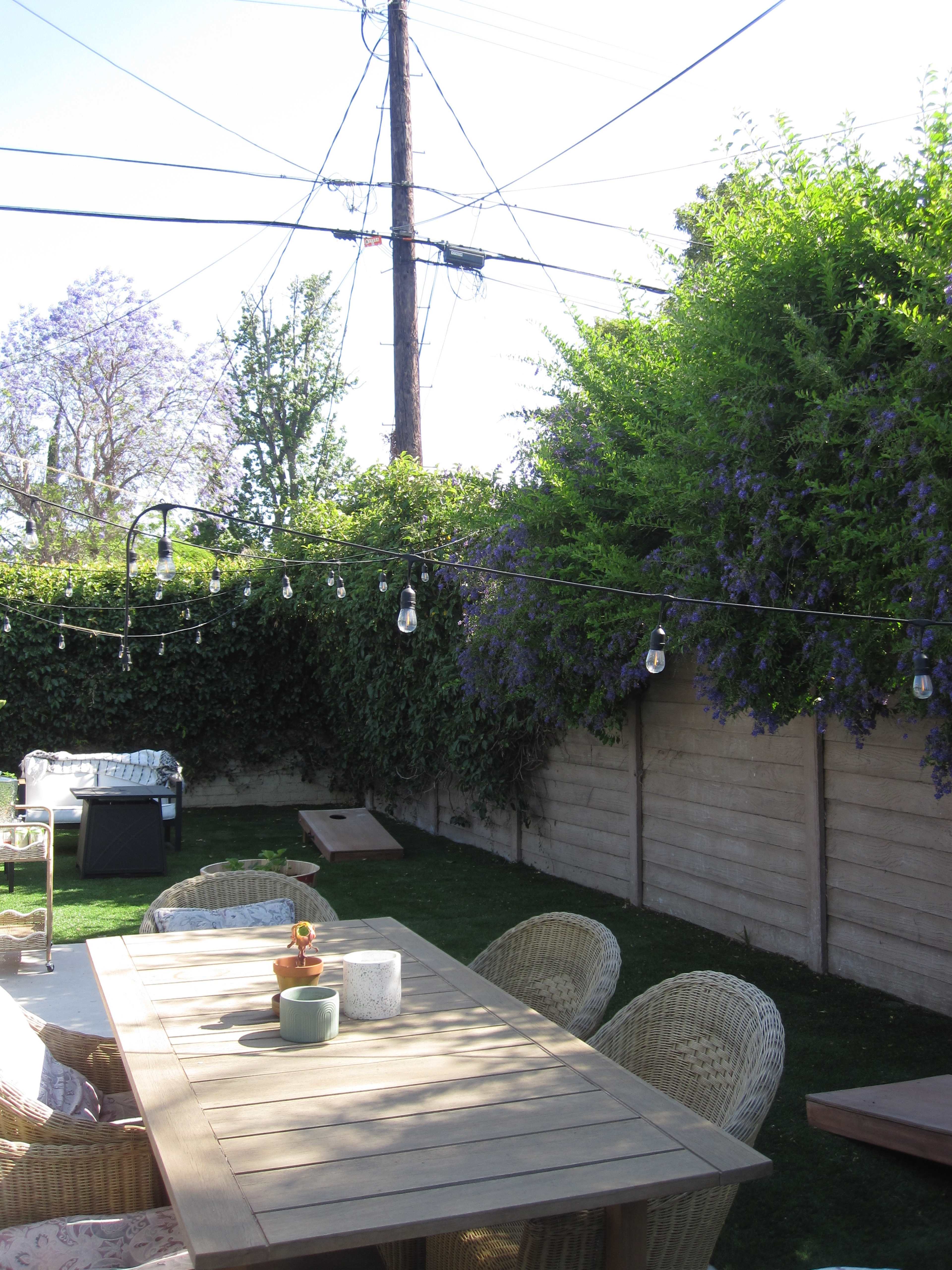 A wooden outdoor table surrounded by wicker chairs is set in a backyard with greenery and string lights overhead.