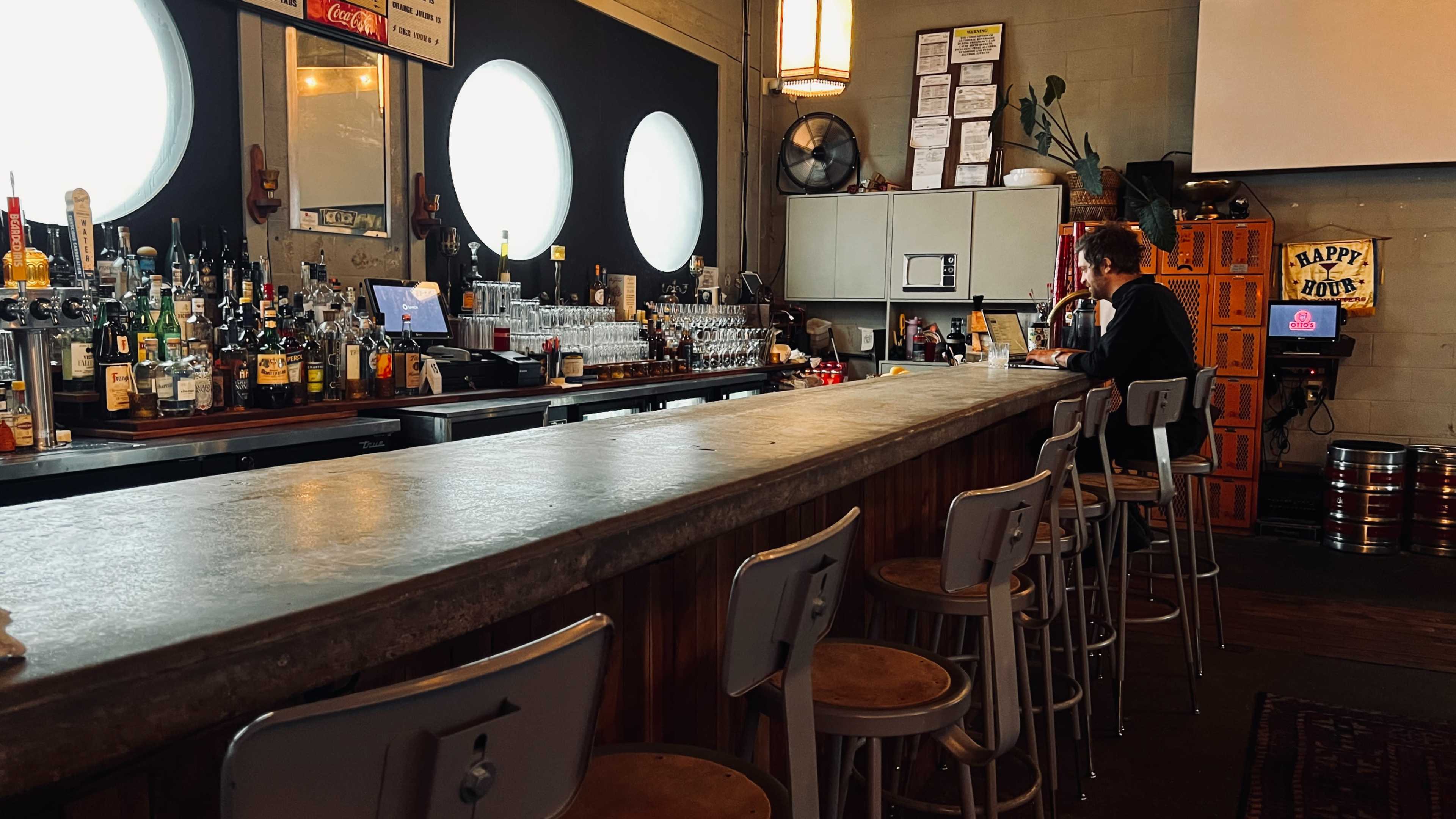 A man sits alone at a long, concrete bar with bottles of various spirits displayed behind him in an establishment with large circular windows.