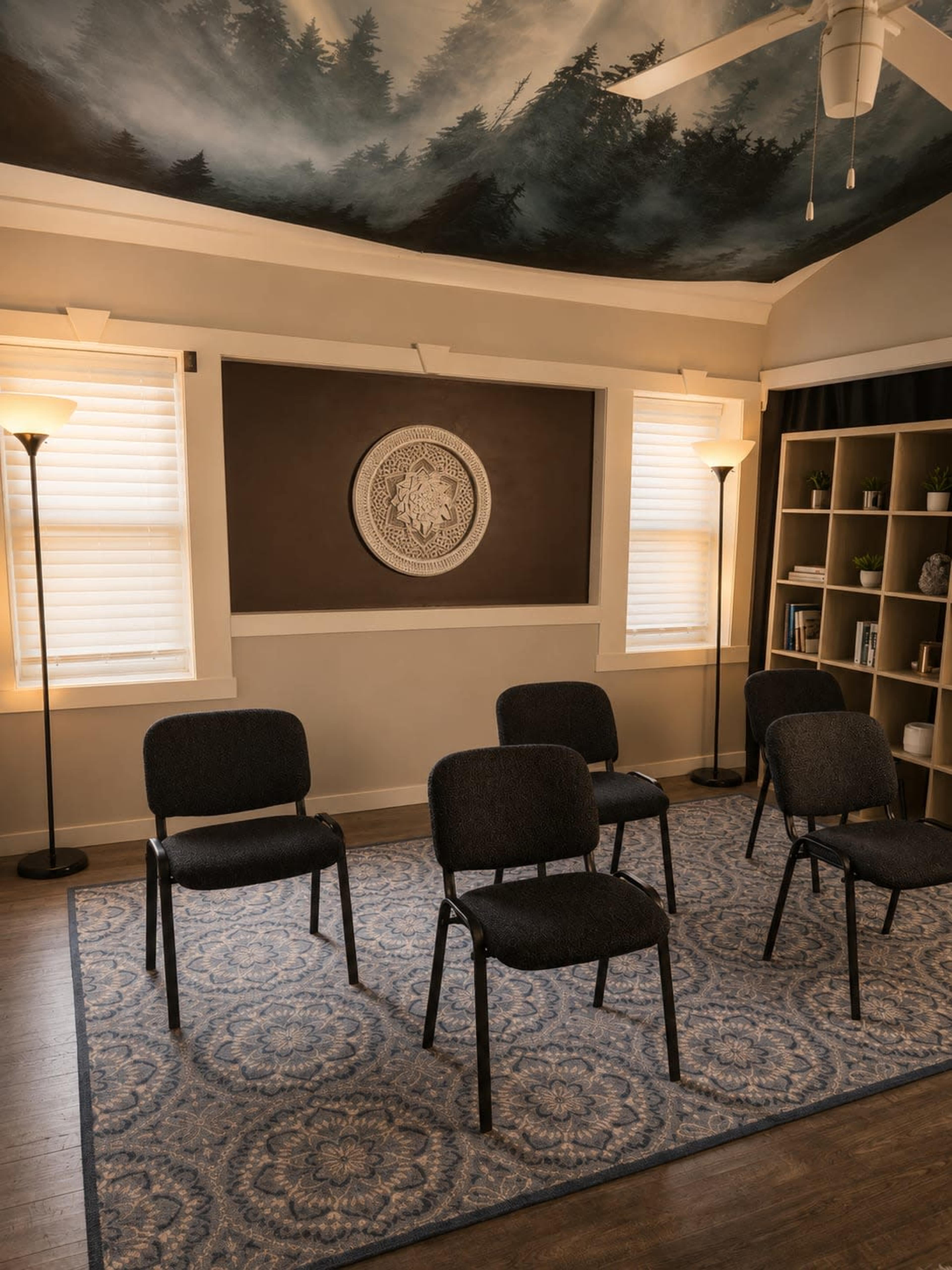 The image shows a room with five empty chairs arranged on a patterned rug, under a mural of trees on the ceiling, with two lamps providing light and a bookshelf against one wall.