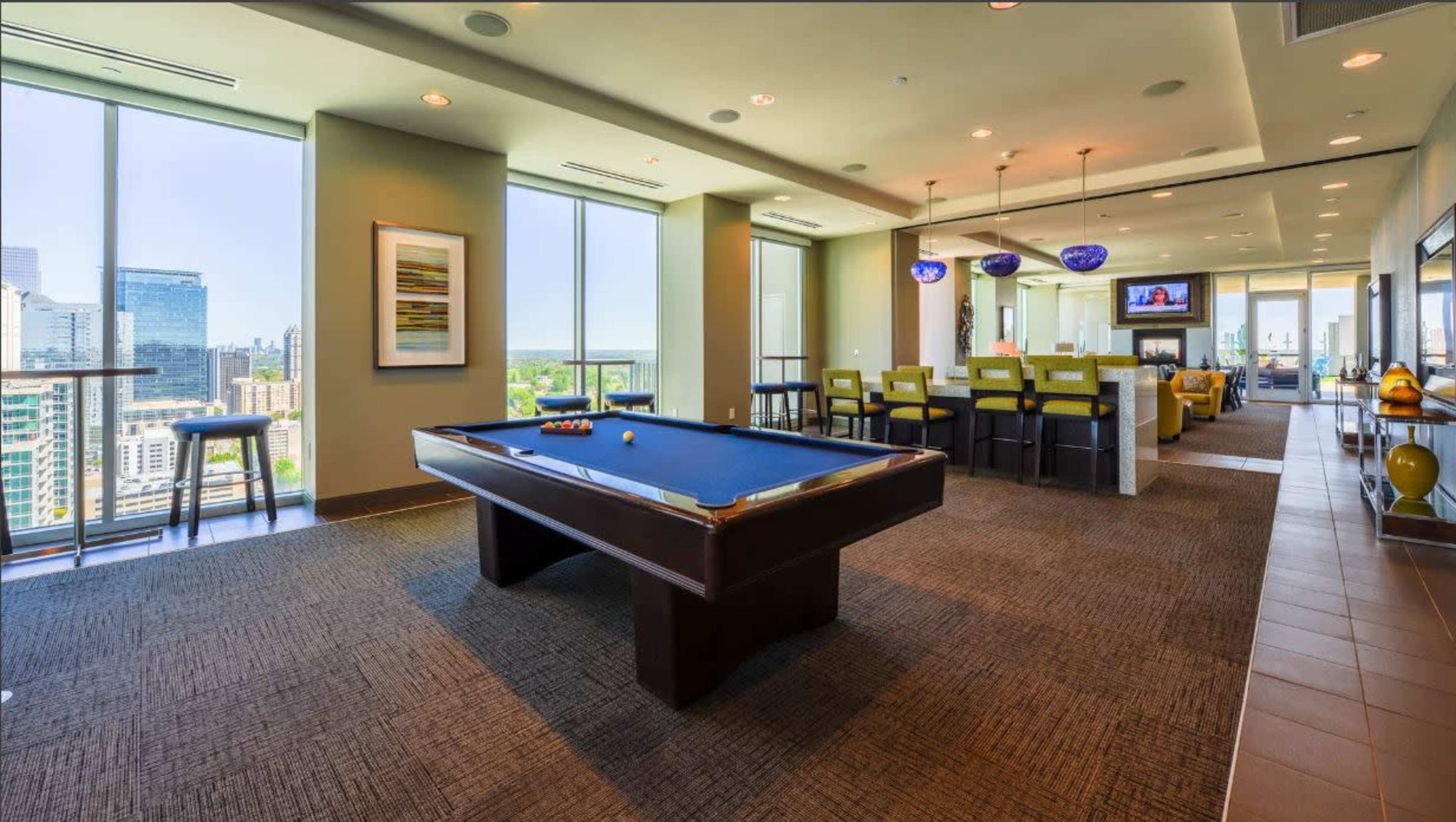 A billiards table is positioned in a well-lit lounge area with large windows showcasing a city view.