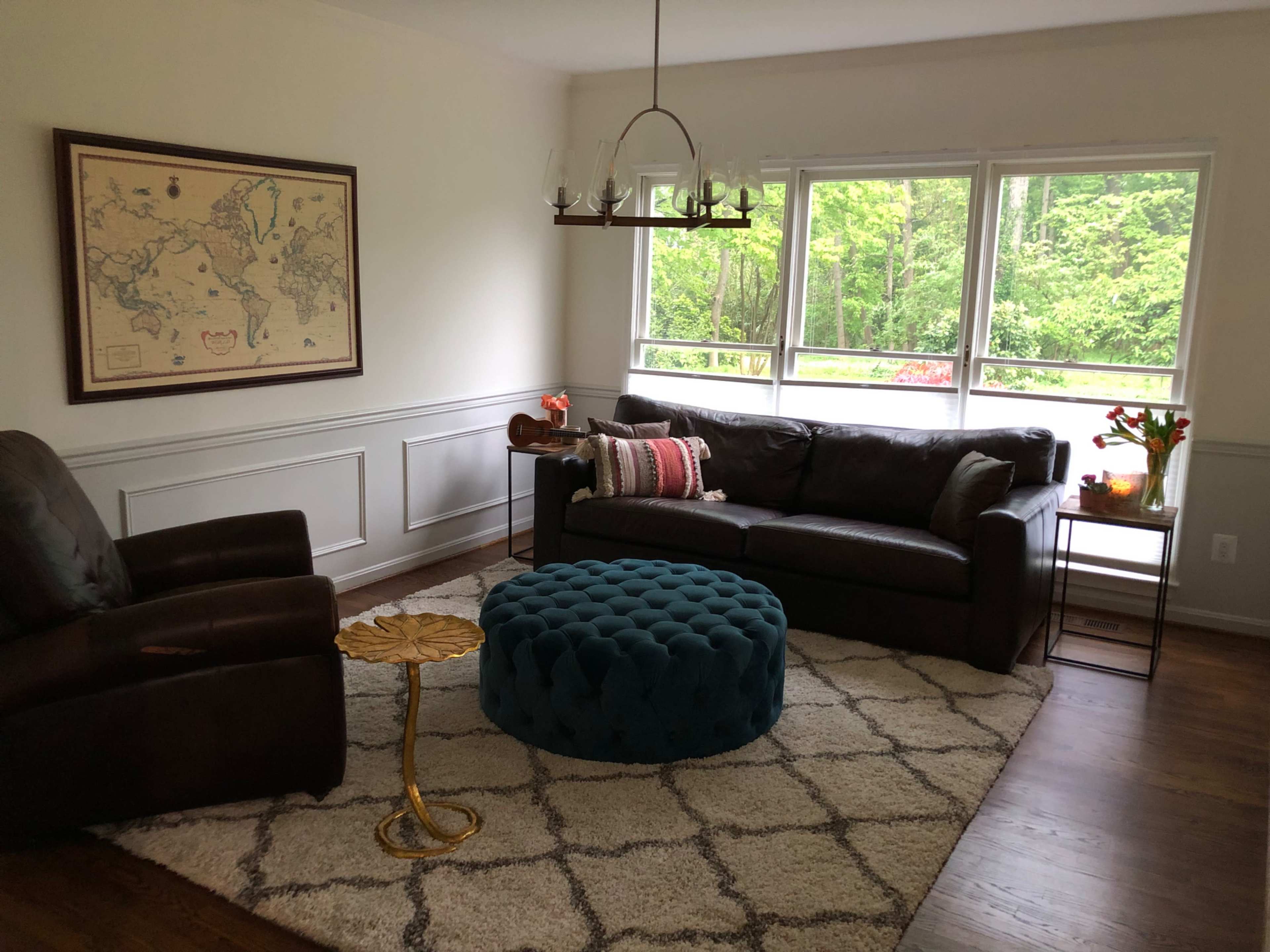 A cozy living room features a brown leather sofa, a blue tufted ottoman, and a vintage world map on the wall, with large windows showcasing a view of greenery outside.