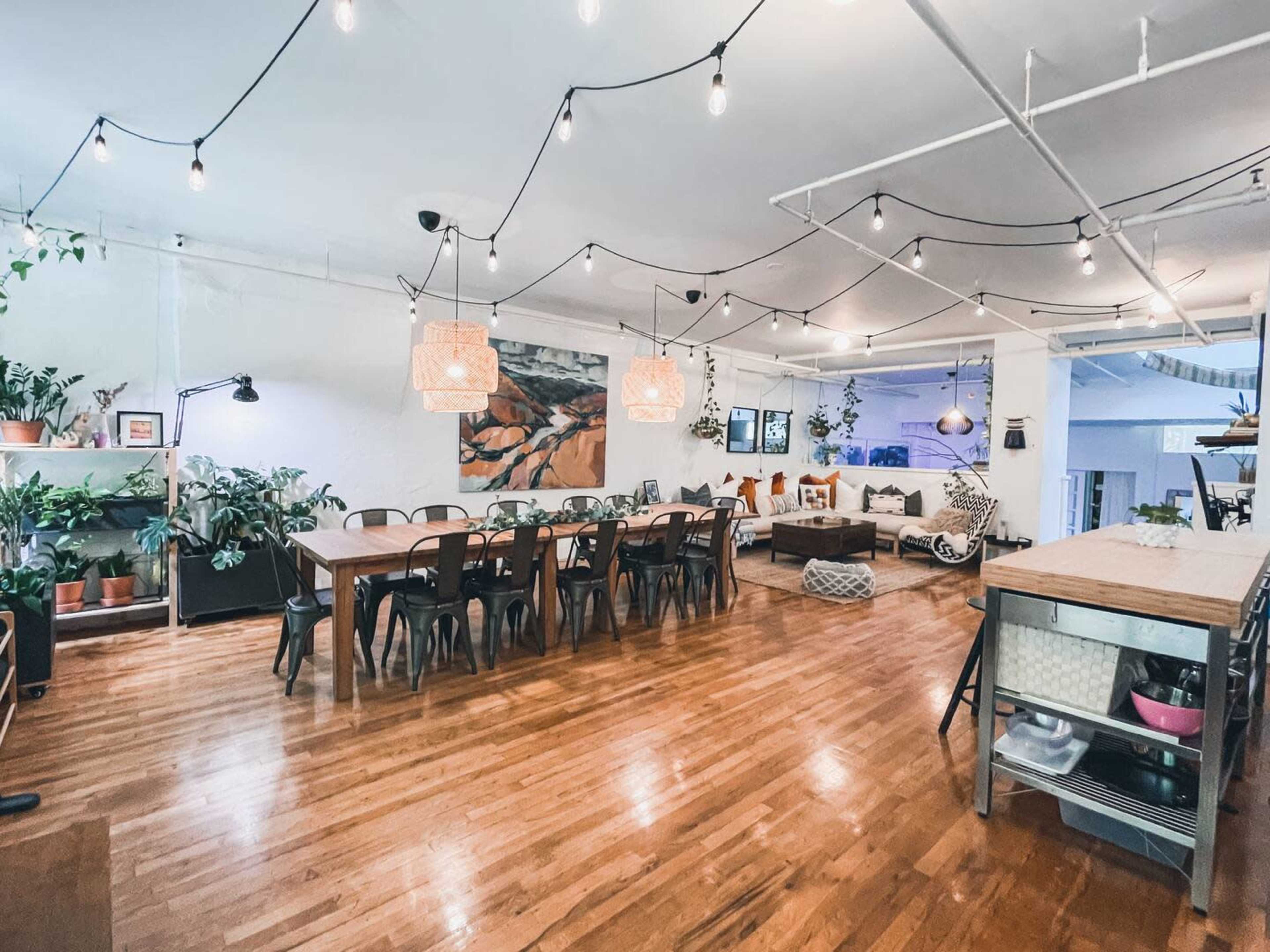 A spacious interior with wooden flooring, a large dining table, string lights overhead, and various plants arranged throughout the room.