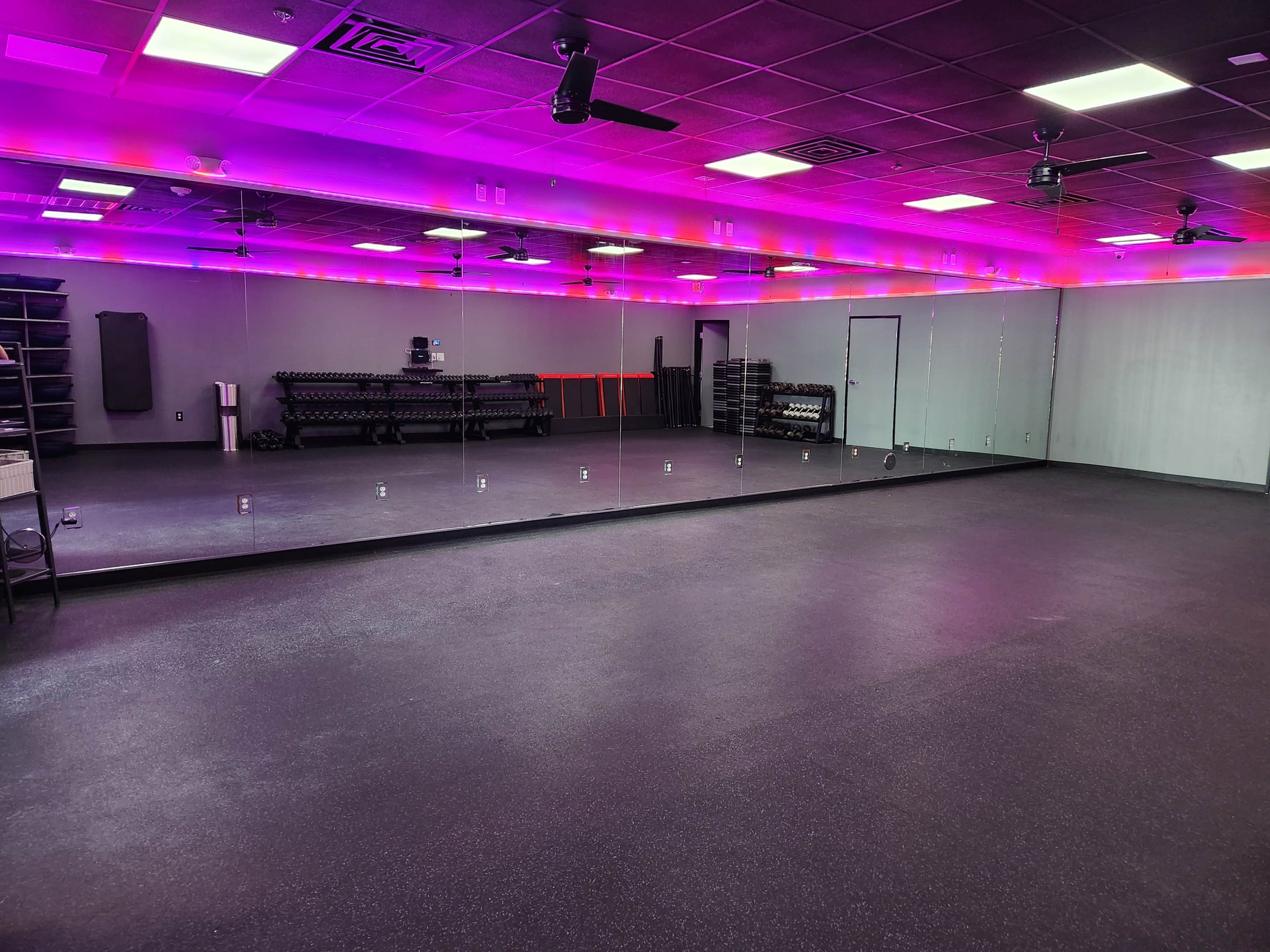 A spacious fitness studio with a large mirrored wall, vibrant lighting, and exercise equipment organized along the sides.