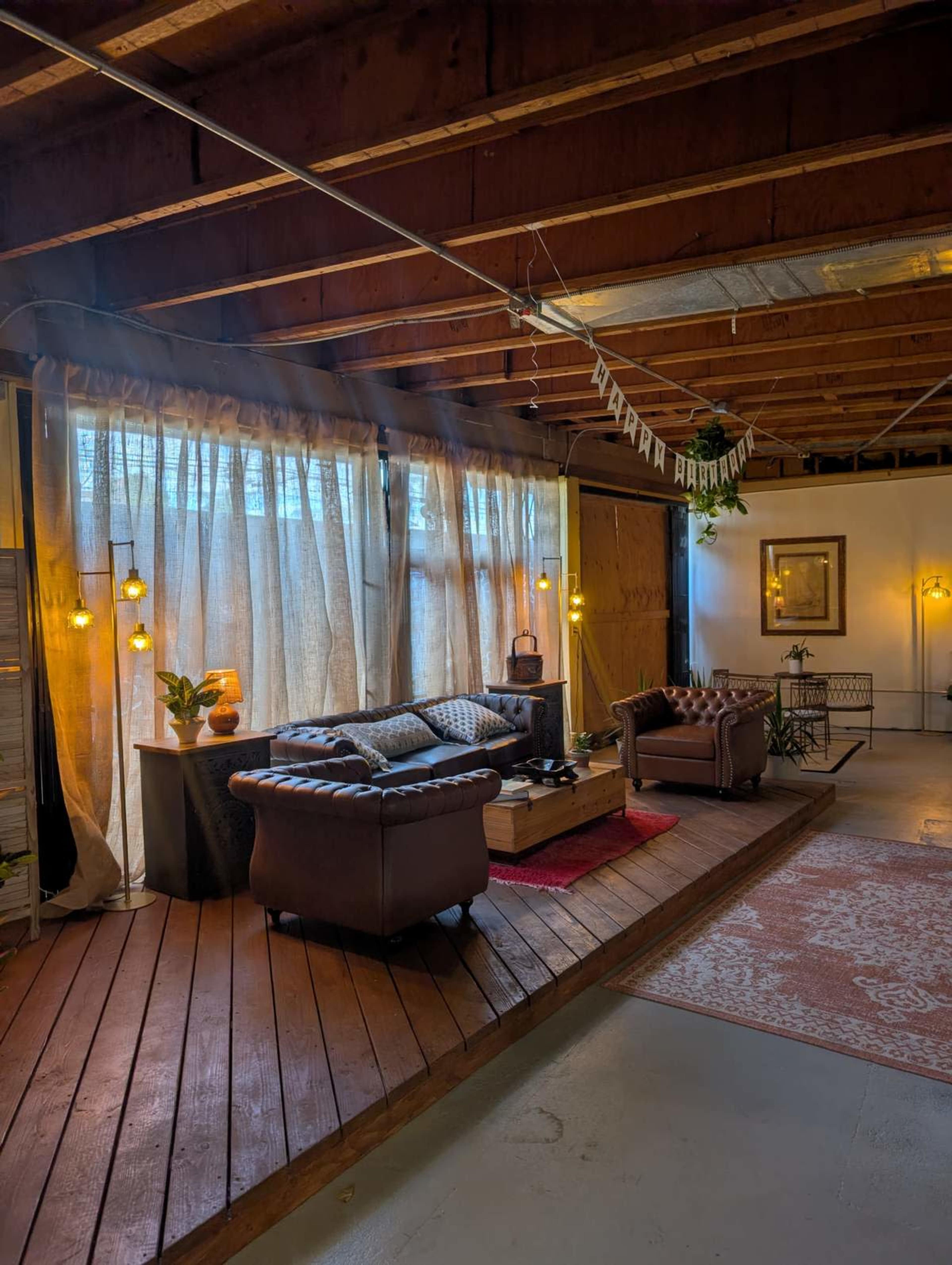 The image shows a cozy living area with a wooden floor, a dark leather couch, large curtains, and various decorative elements.