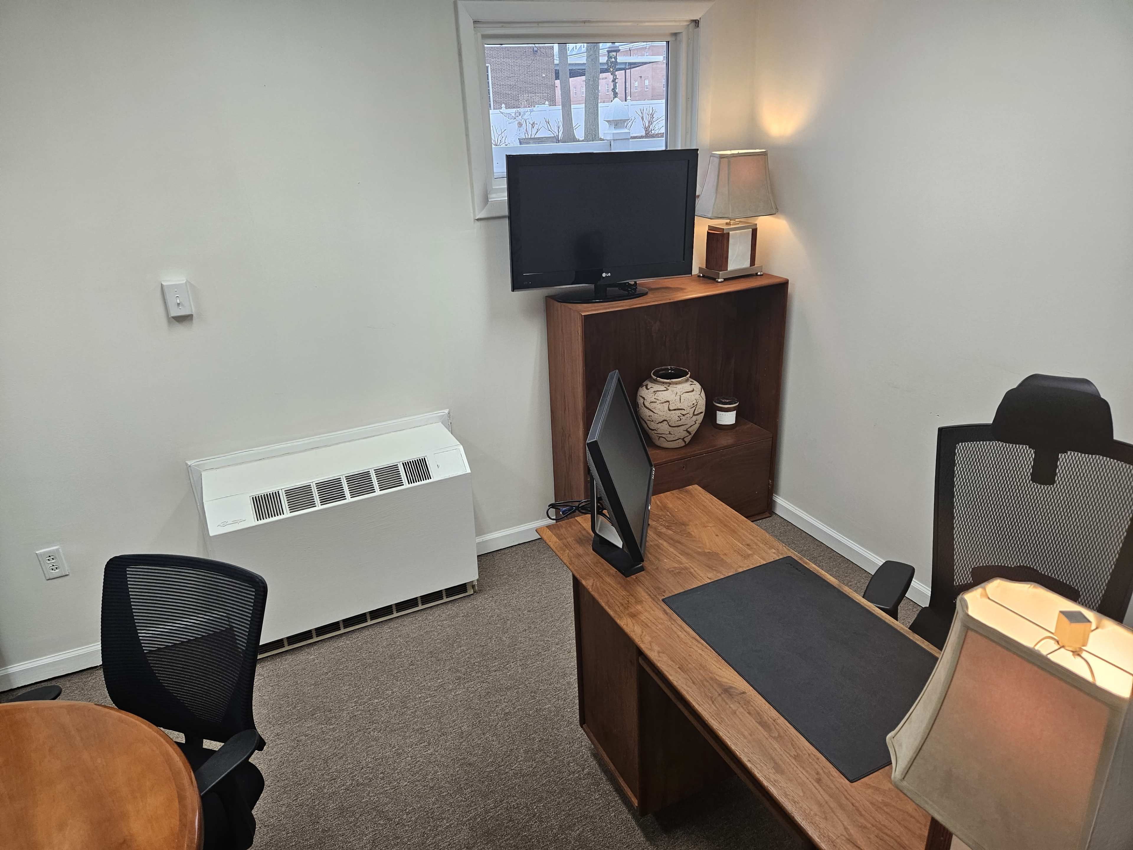 The image shows a small office space with a wooden desk, a computer monitor, a television on a cabinet, and two chairs, all illuminated by natural light from a window.