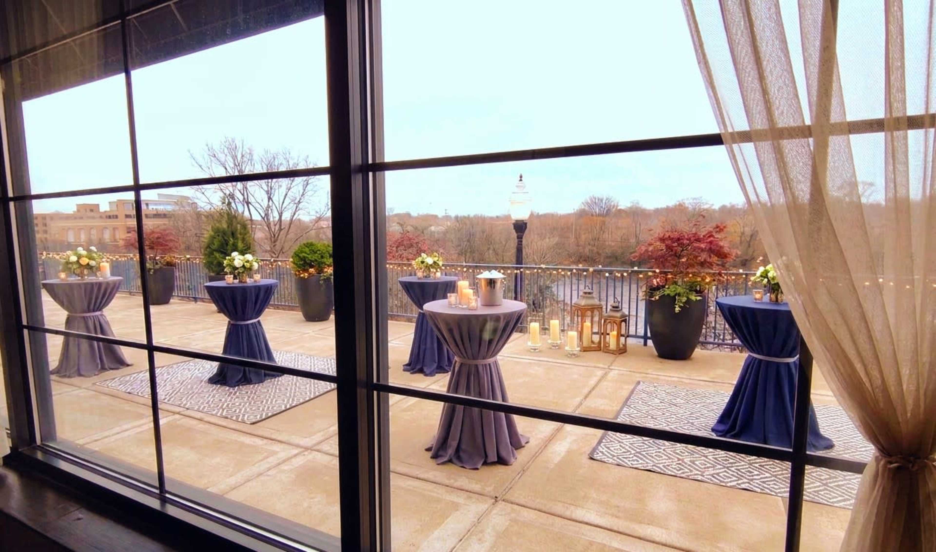 The image shows a venue patio with tables dressed in dark blue linen, arranged on a concrete surface, overlooking a river with decorative candles and flowers.