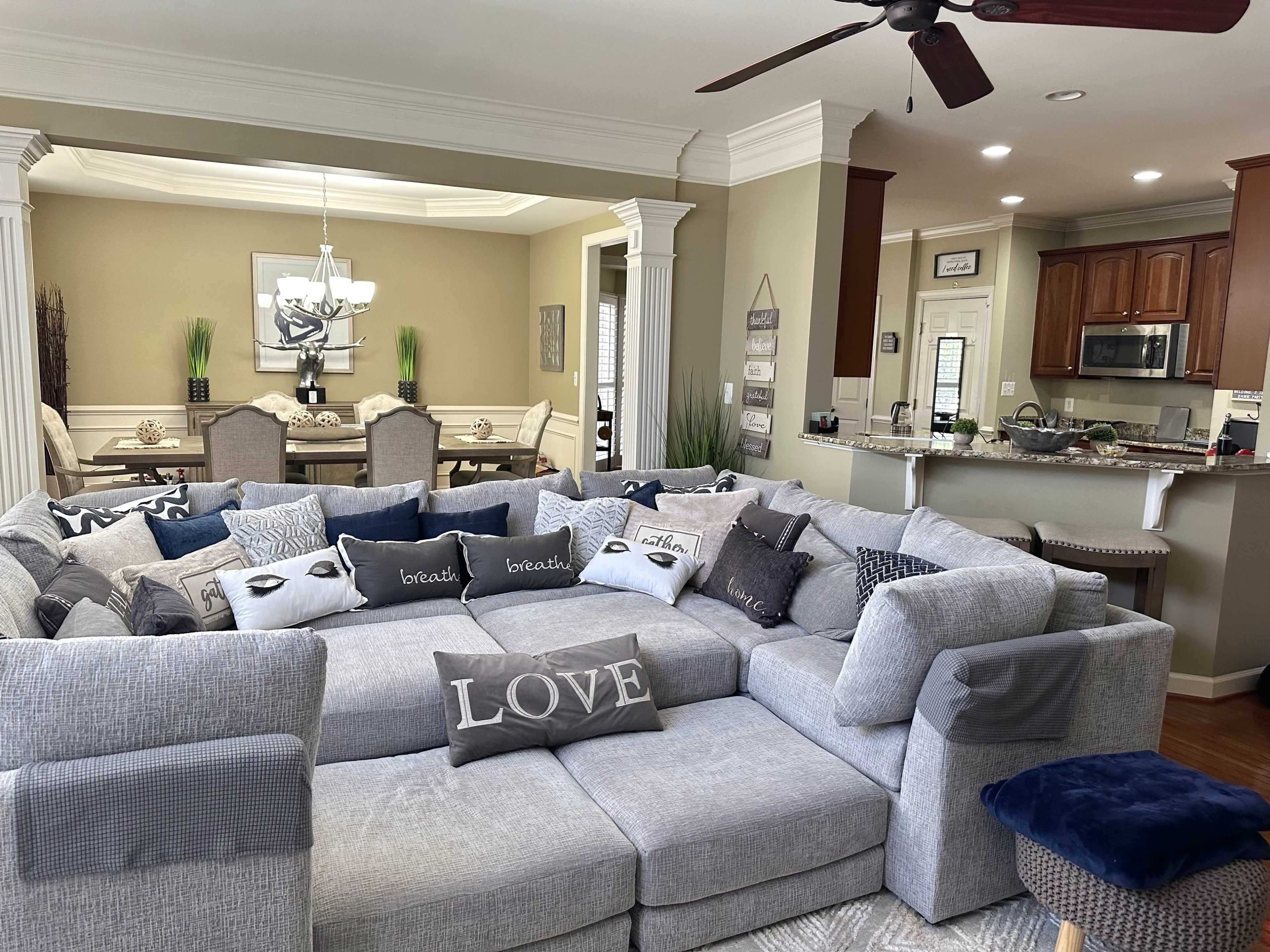 The image features a spacious living room with a large sectional sofa arranged in a U-shape, adorned with decorative pillows, overlooking a dining area with a table and chairs.